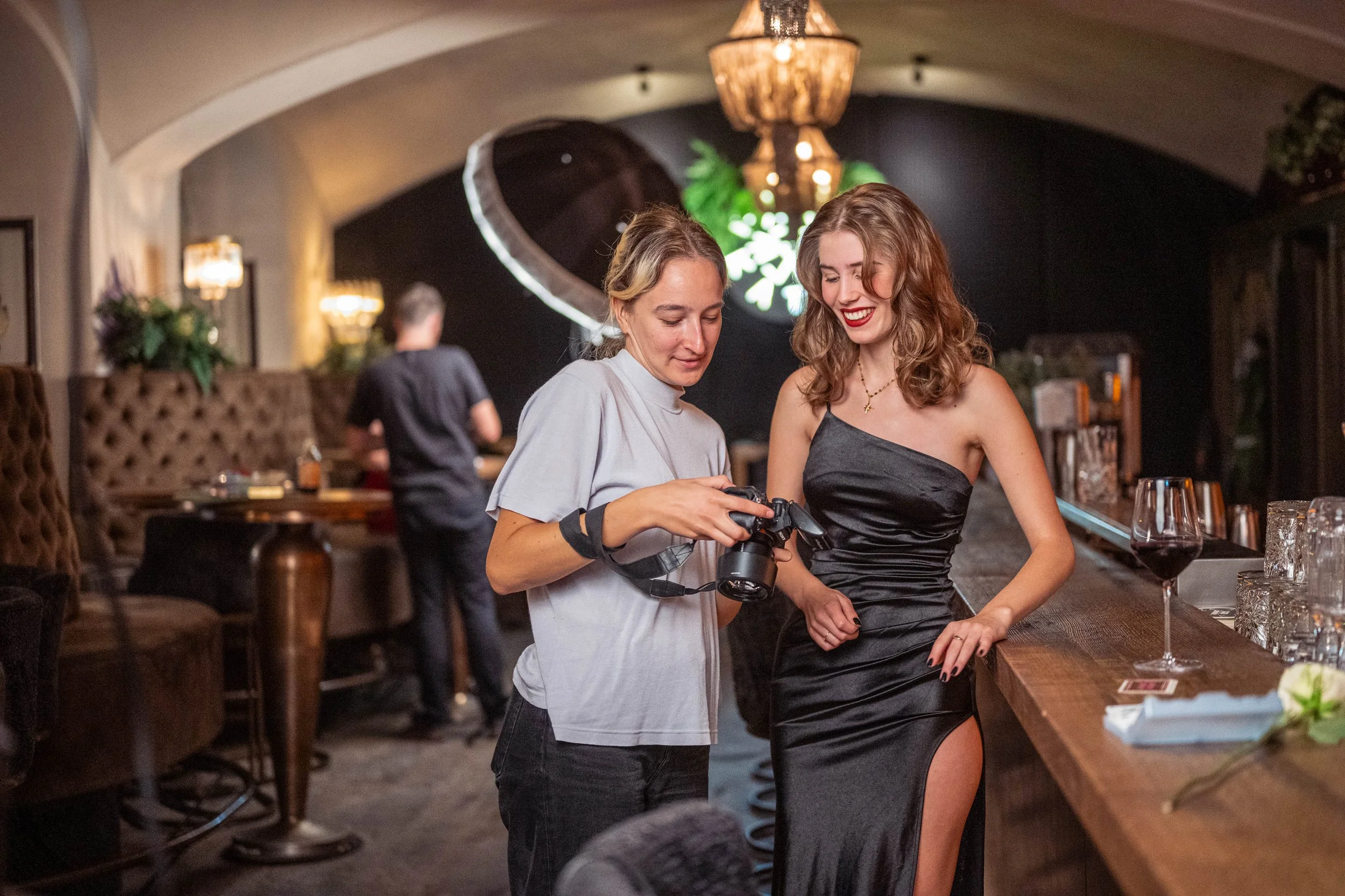 Zwei Frauen, eine mit einem schwarzen Abendkleid und eine mit einem weißen T-Shirt, schauen sich Fotos auf einer Kamera an, in einem eleganten Restaurant oder Bar, mit Wein und Dekorationen im Hintergrund.
