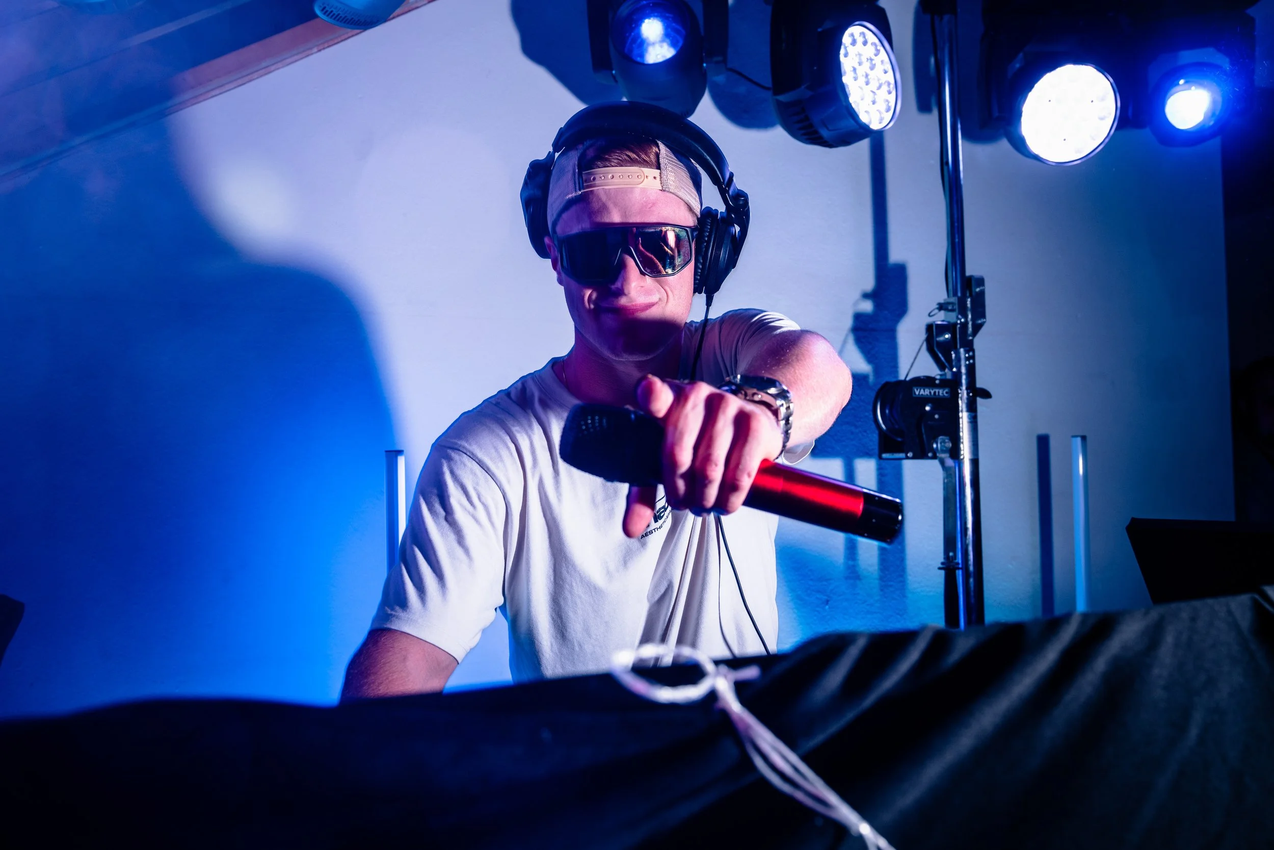DJ bei einer Live-Performance mit Sonnenbrille, Kabel 10 Uhren, Transparentem Baseball Cap, auf einem Club- oder Festival-Bühne, das Licht ist blau.