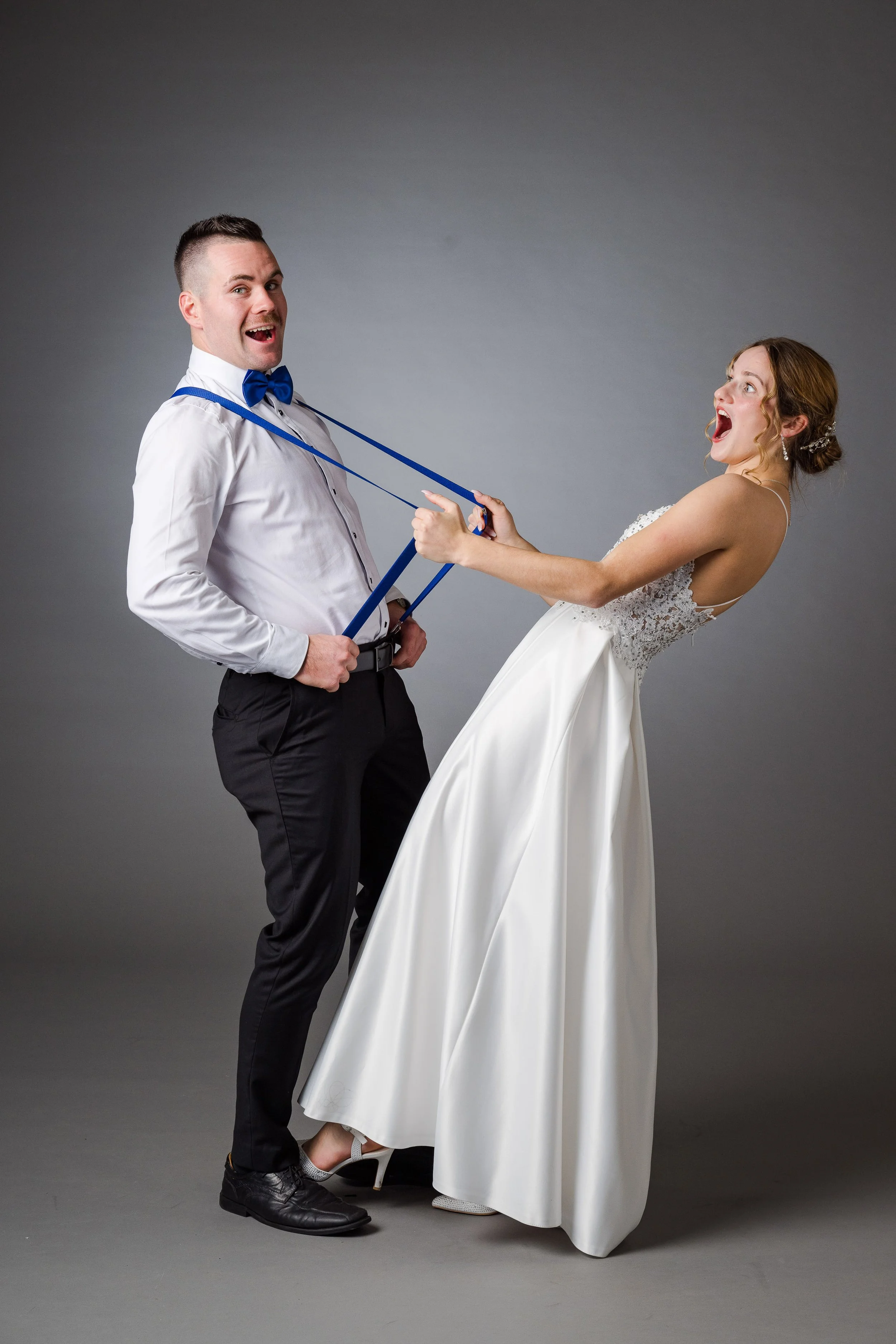 Ein Brautpaar spielt mit einem Blauer Bindegürtel bei einem Fotoshooting vor einem grauen Hintergrund.