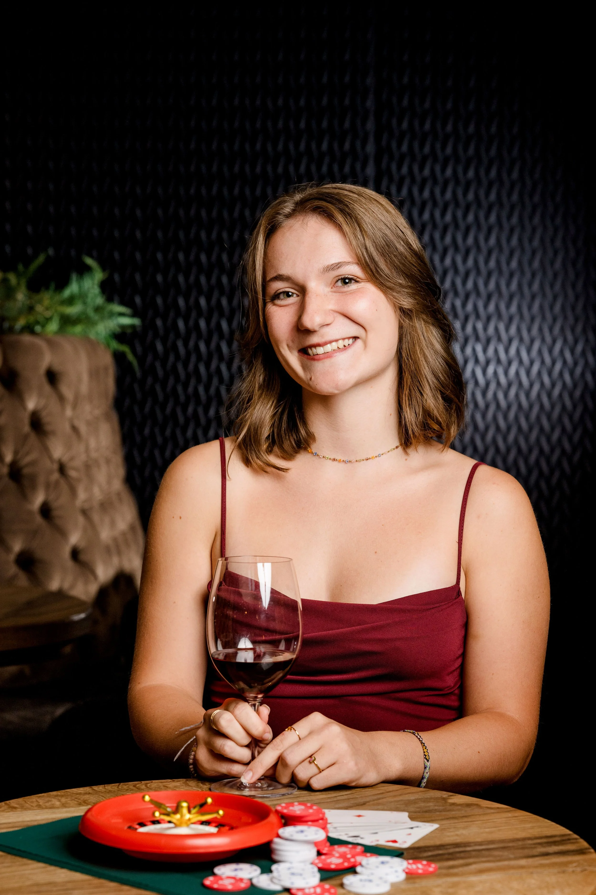 Eine junge Frau mit braunen, mittellangen Haaren, die in einem roten Träger-Top mit einer Glas Rotwein in der Hand lächelt. Sie sitzt an einem Tisch mit verschiedenen Pokerchips, Karten und einem roten Poker-Table-Automaten. Hinter ihr eine dunkle, s