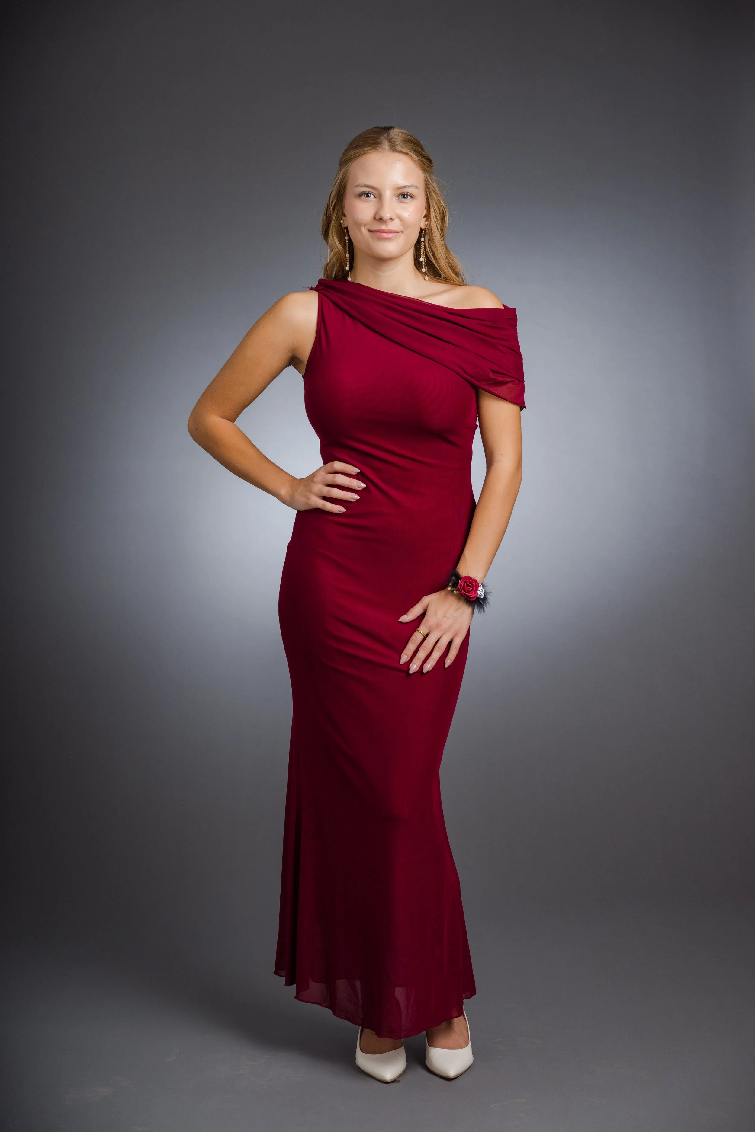 Eine junge Frau in einem roten, bodenlangen Abendkleid mit Off-Shoulder-Design steht vor grauem Hintergrund.