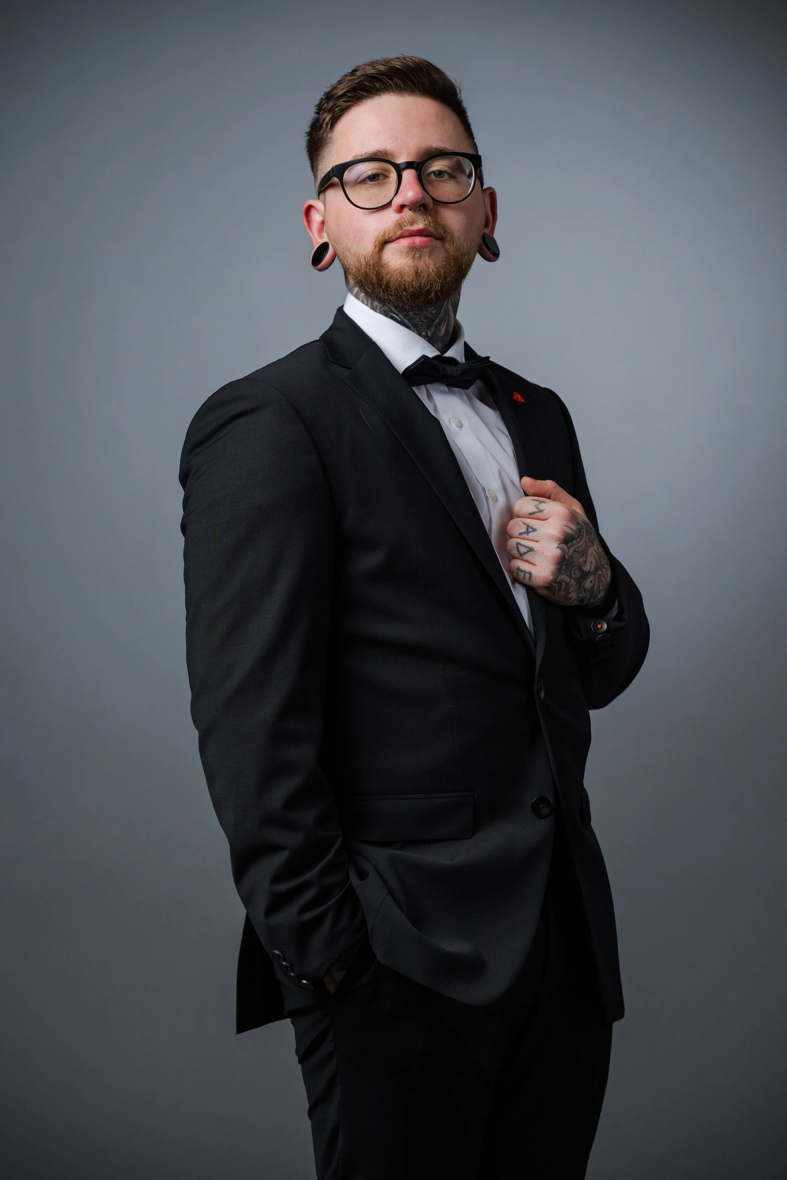 Ein junger Mann in einem schwarzen Anzug mit weißen Hemd und Fliege steht vor grauem Hintergrund. Er trägt Brille, hat Tattoos am Hals und Hände, sowie große Ohrpiercings.