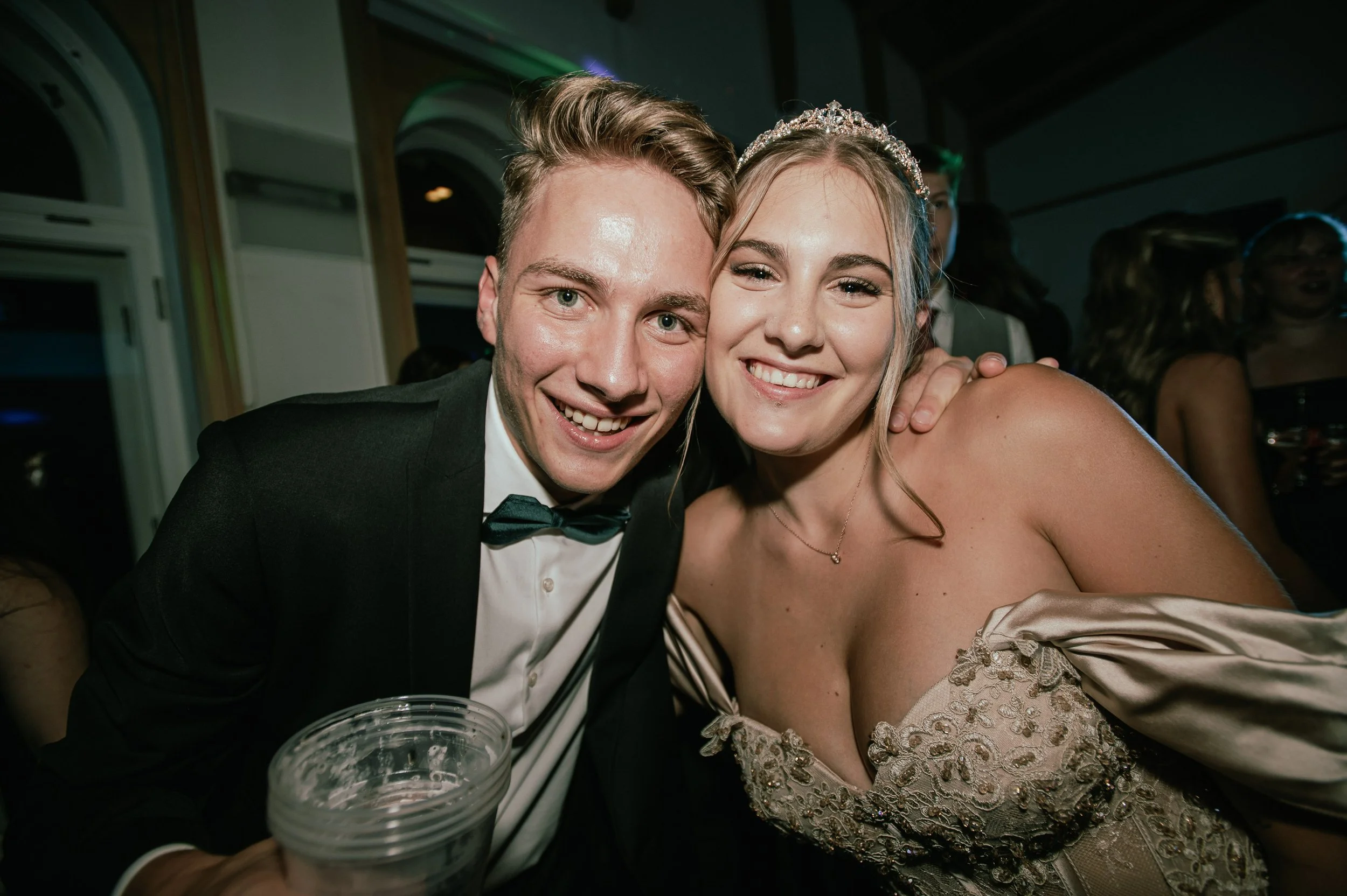 Ein glückliches Paar bei einer Feier, vermutlich einem Ball oder einer Hochzeit, beide lächelnd in festlicher Kleidung, der Mann trägt einen schwarzen Anzug mit Fliege und die Frau ein elegantes, cremefarbendes Kleid mit Verzierungen und einer Tiara.