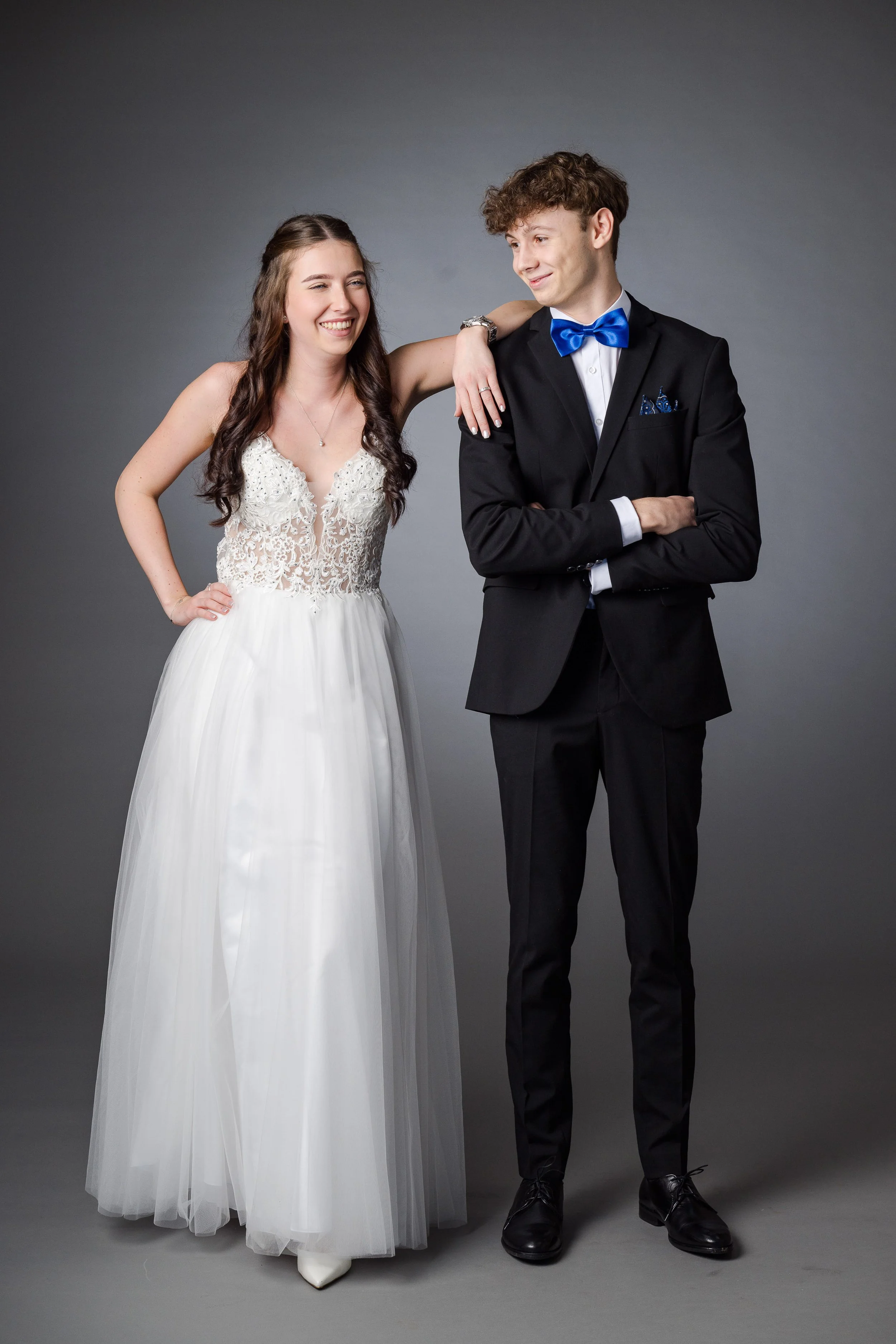 Ein Brautpaar in Hochzeitskleidung posiert vor einem grauen Hintergrund. Die Braut trägt ein weißes Brautkleid mit Spitze und die Haare sind lang und lockig. Der Bräutigam trägt einen schwarzen Anzug mit weißem Hemd, blauer Fliege und einem blauen Ei