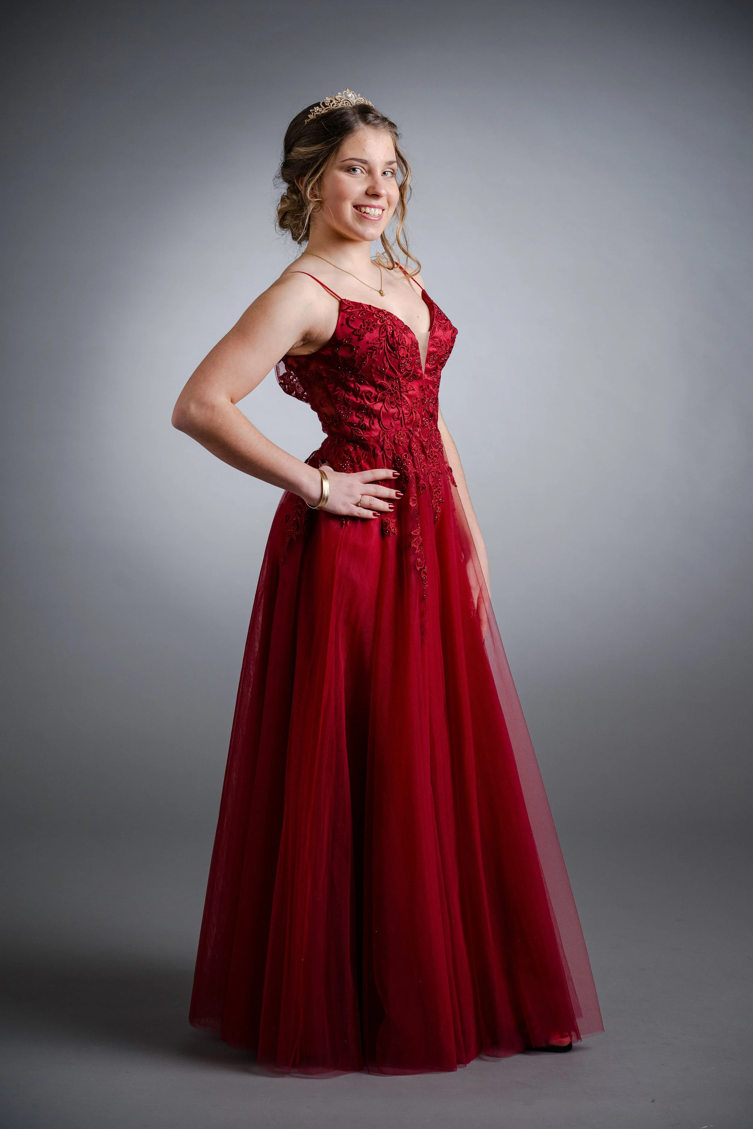 Junge Frau in rotem Brautkleid mit Tiara und Goldschmuck, lächelnd, vor grauem Hintergrund.
