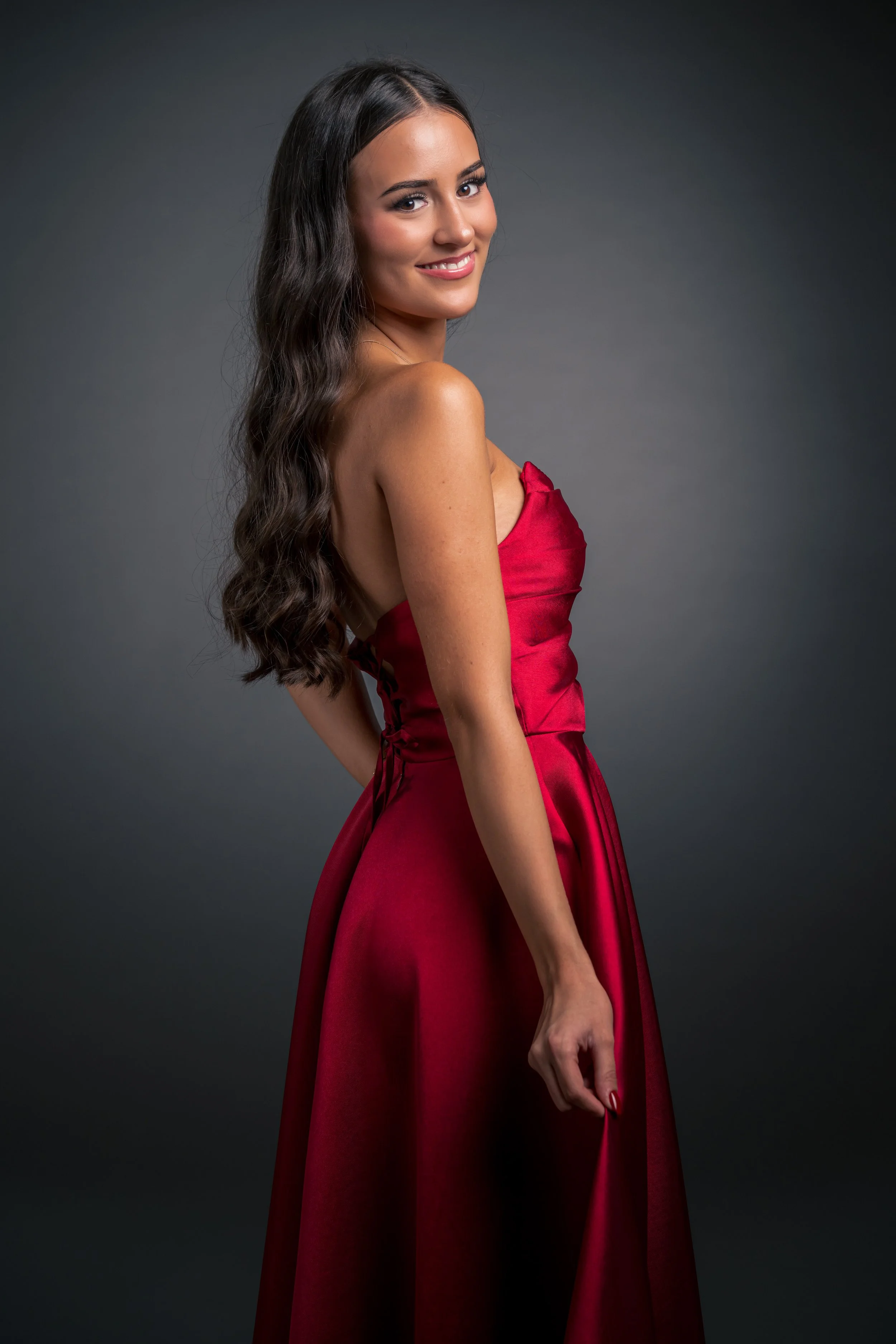 Eine junge Frau trägt ein rotes, elegantes Abendkleid, hat lange, lockige dunkle Haare und lächelt in die Kamera.