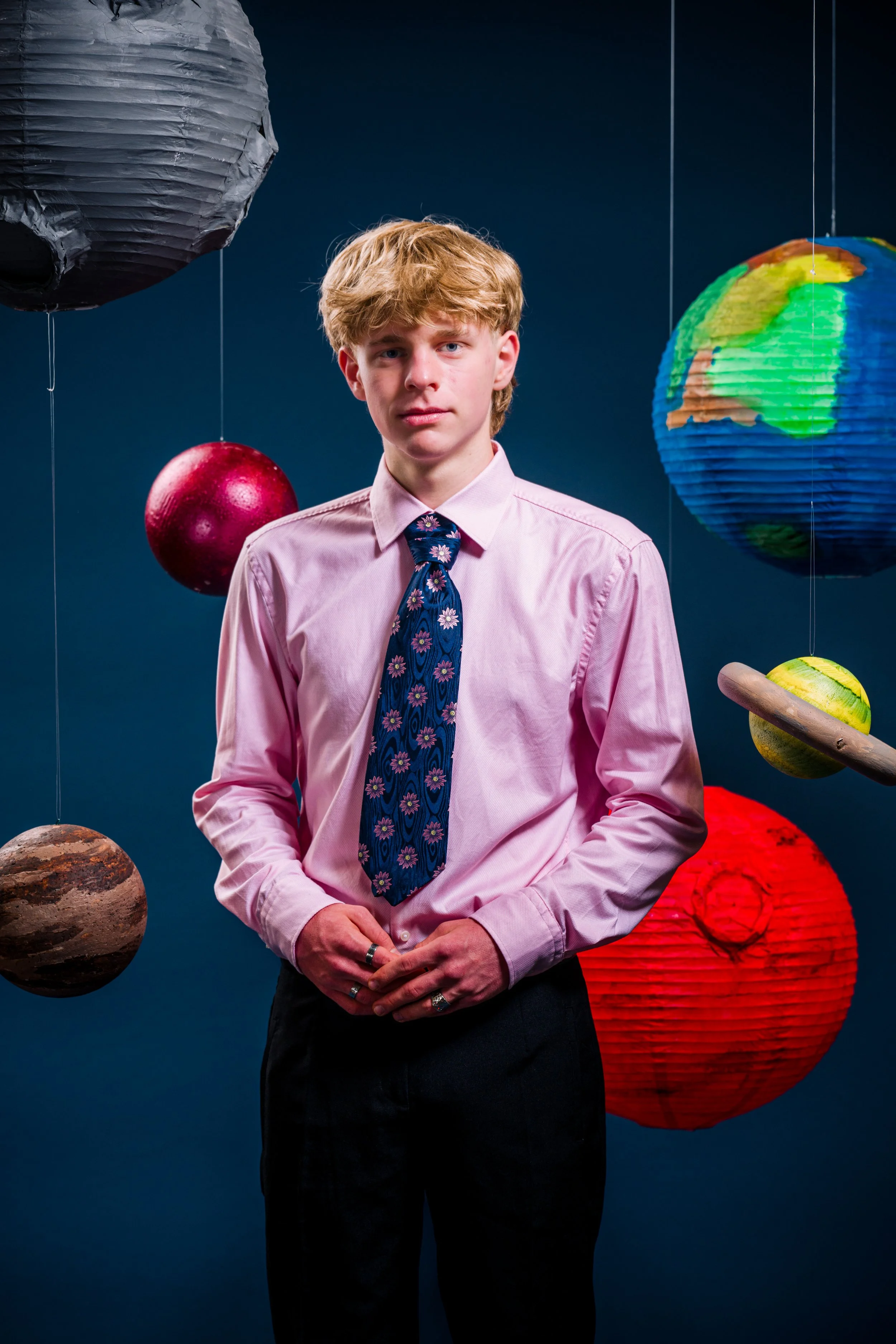 Ein junger Mann trägt ein rosa Hemd mit einer blumigen Krawatte, umgeben von schwebenden, bunten Laternen in Form von Planeten und der Erde.