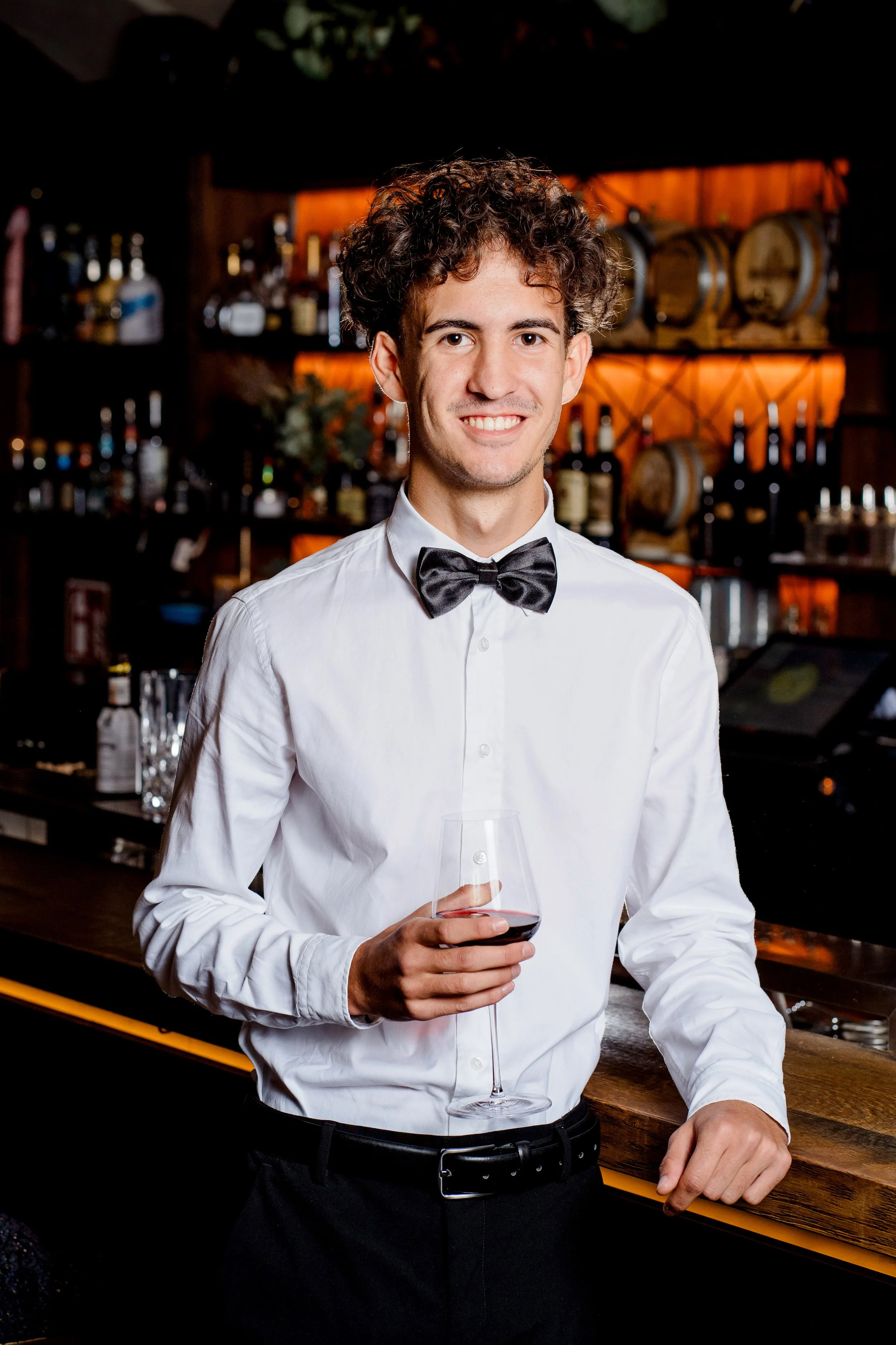 Ein junger Mann im weißen Hemd mit Fliege steht in einer Bar, hält ein Glas Rotwein und lächelt in die Kamera.