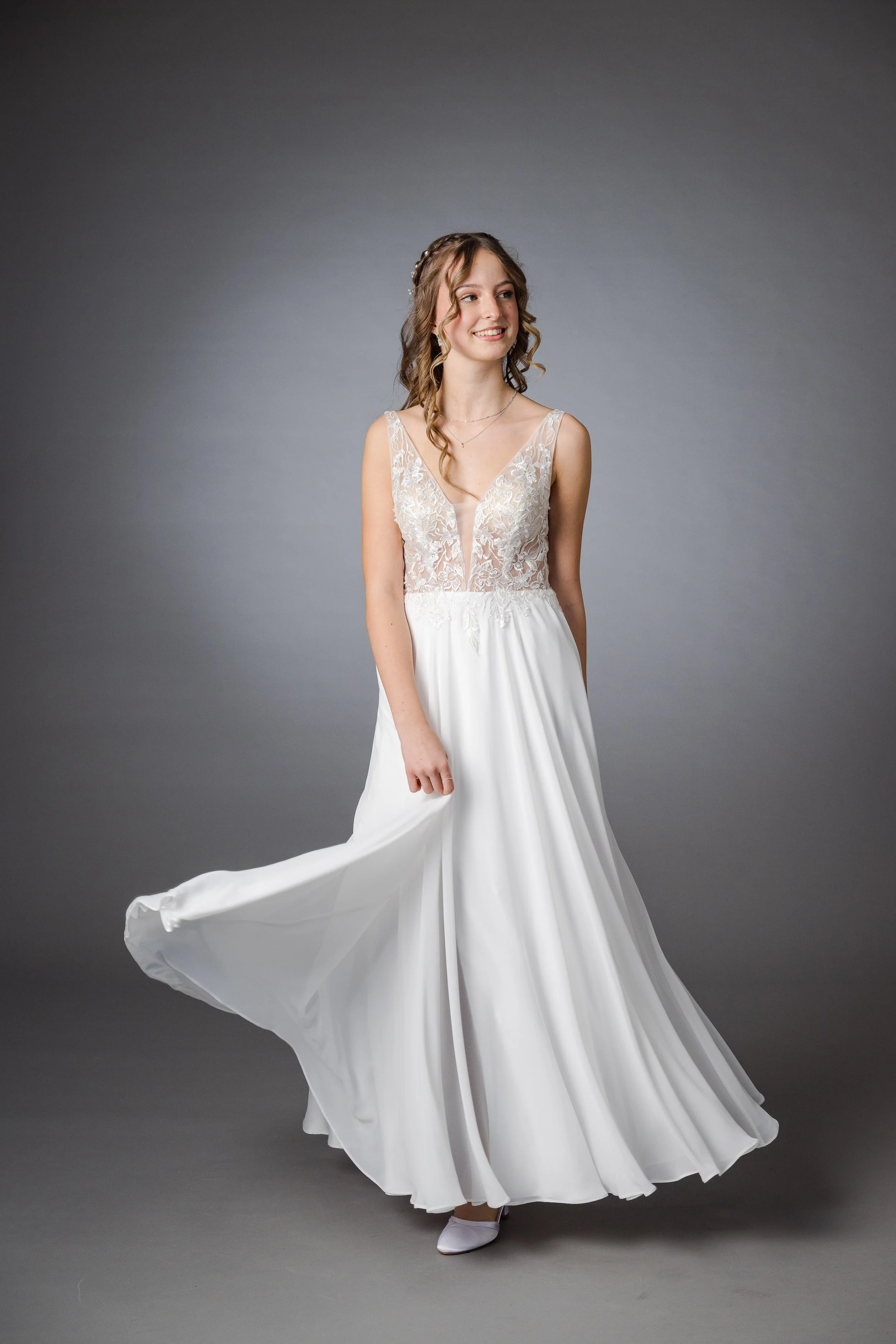 Junge Frau in einem weißen Hochzeitskleid mit Spitzen-Top, die vor einem grauen Hintergrund steht und lächelt.
