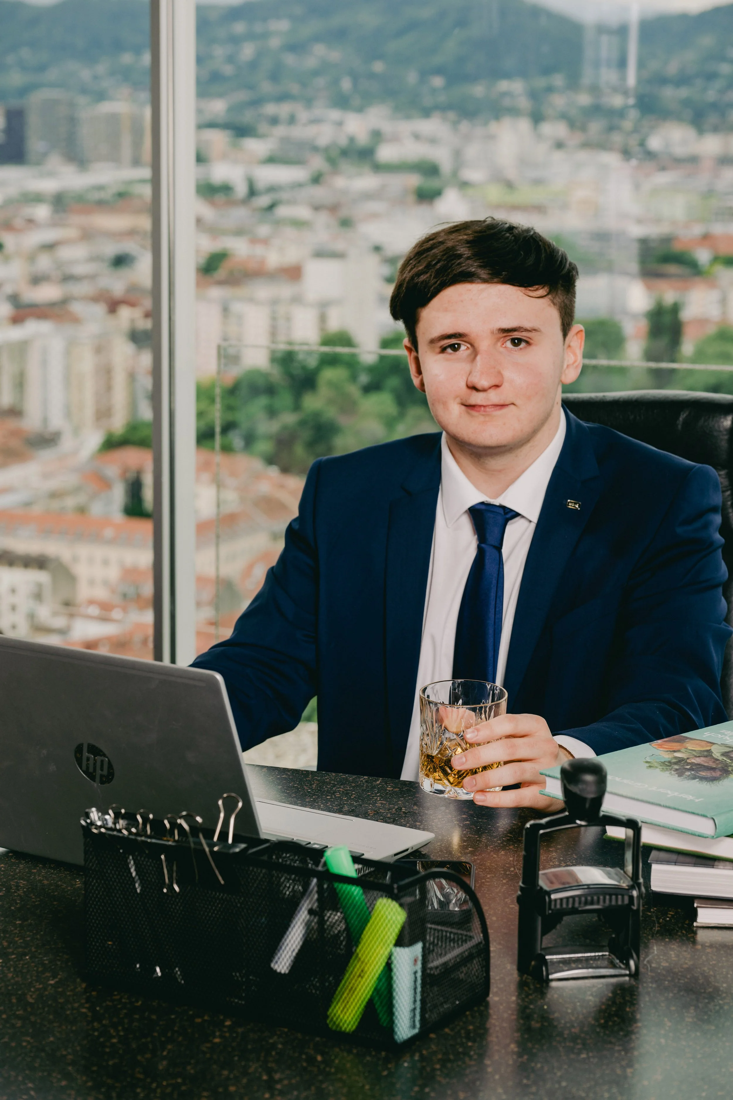Junger Mann in Anzug sitzt am Schreibtisch im Büro mit Blick auf eine Stadt, hält ein Glas Whisky in der Hand, Laptop, Stempel, Büroklammern und Buch auf dem Schreibtisch.