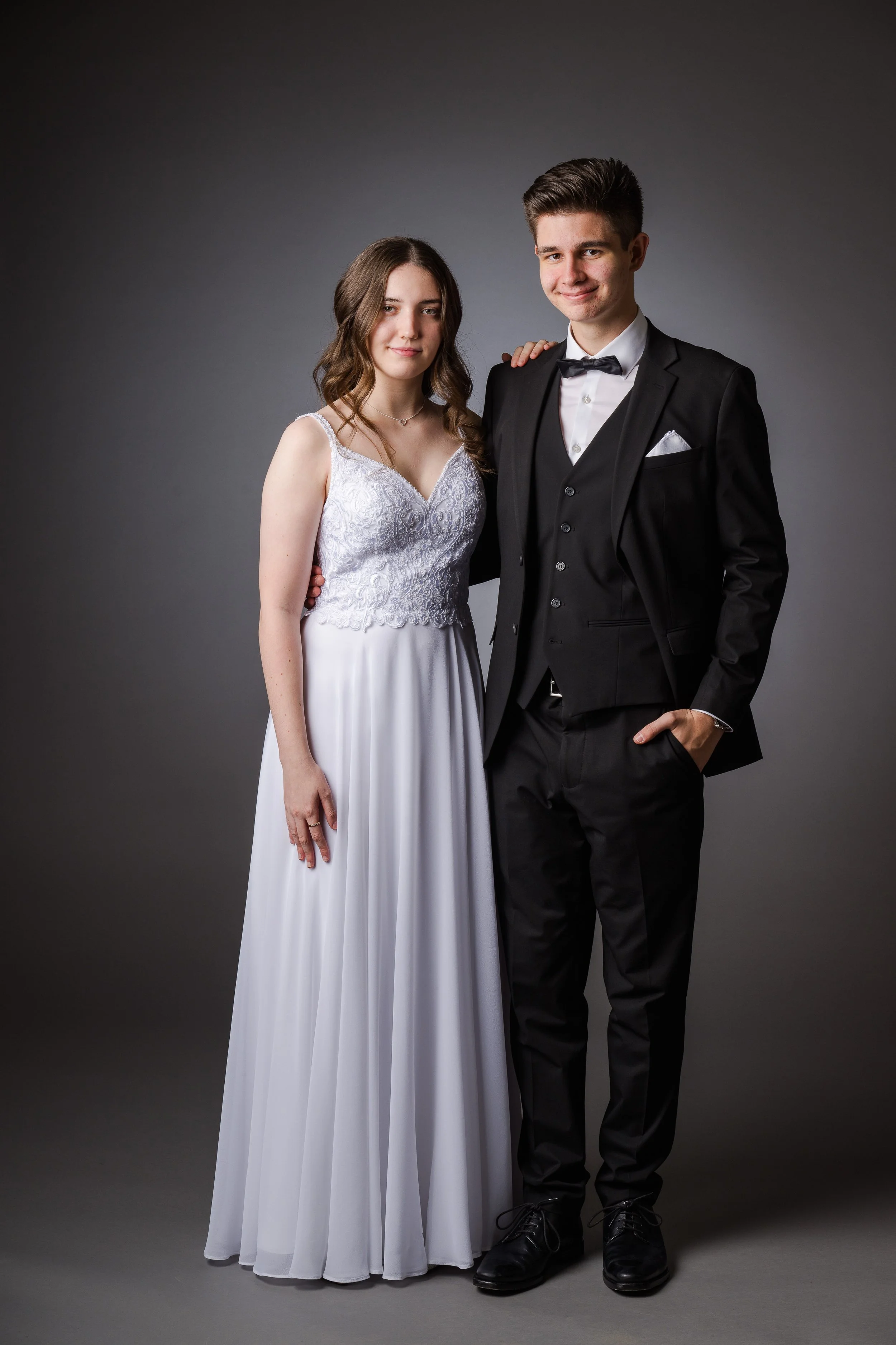 Ein junges Paar in formeller Kleidung, vermutlich für eine besondere Gelegenheit wie eine Hochzeit, vor einem grauen Hintergrund. Die Frau trägt ein weißes Kleid mit Stickerei am Oberteil, der Mann trägt einen schwarzen Anzug mit Fliege.