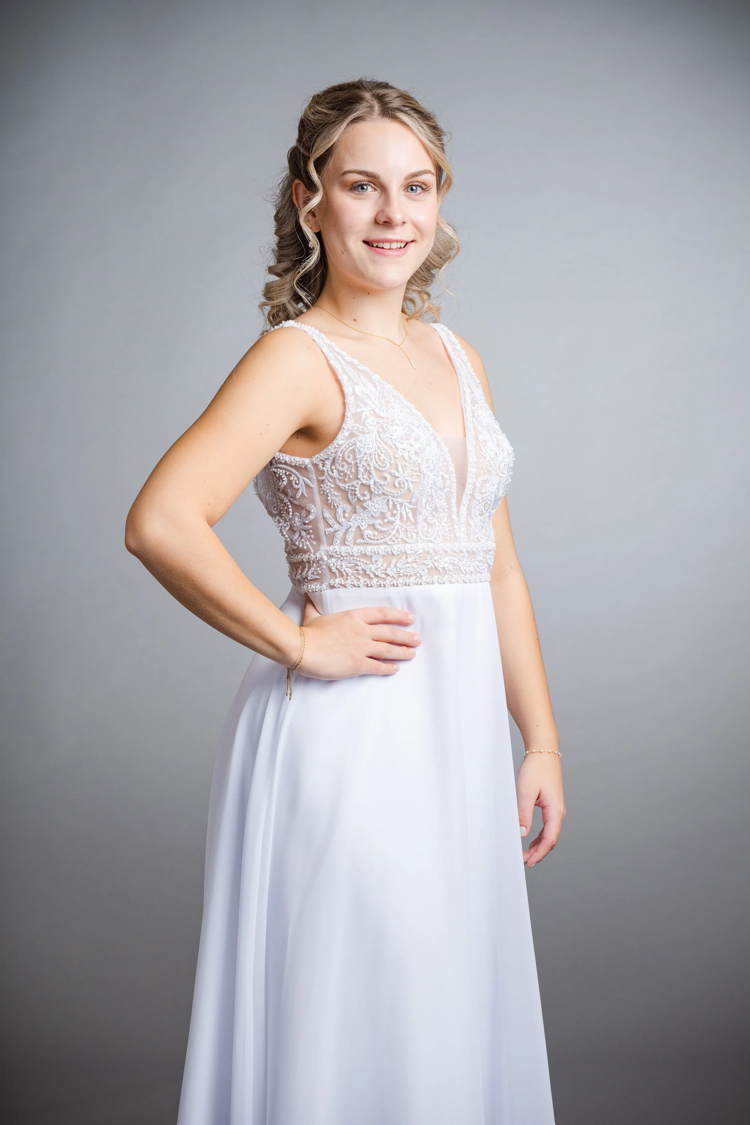 Eine junge Frau in einem weißen Brautkleid mit stickerten Trägern steht vor einem grauen Hintergrund und lächelt.