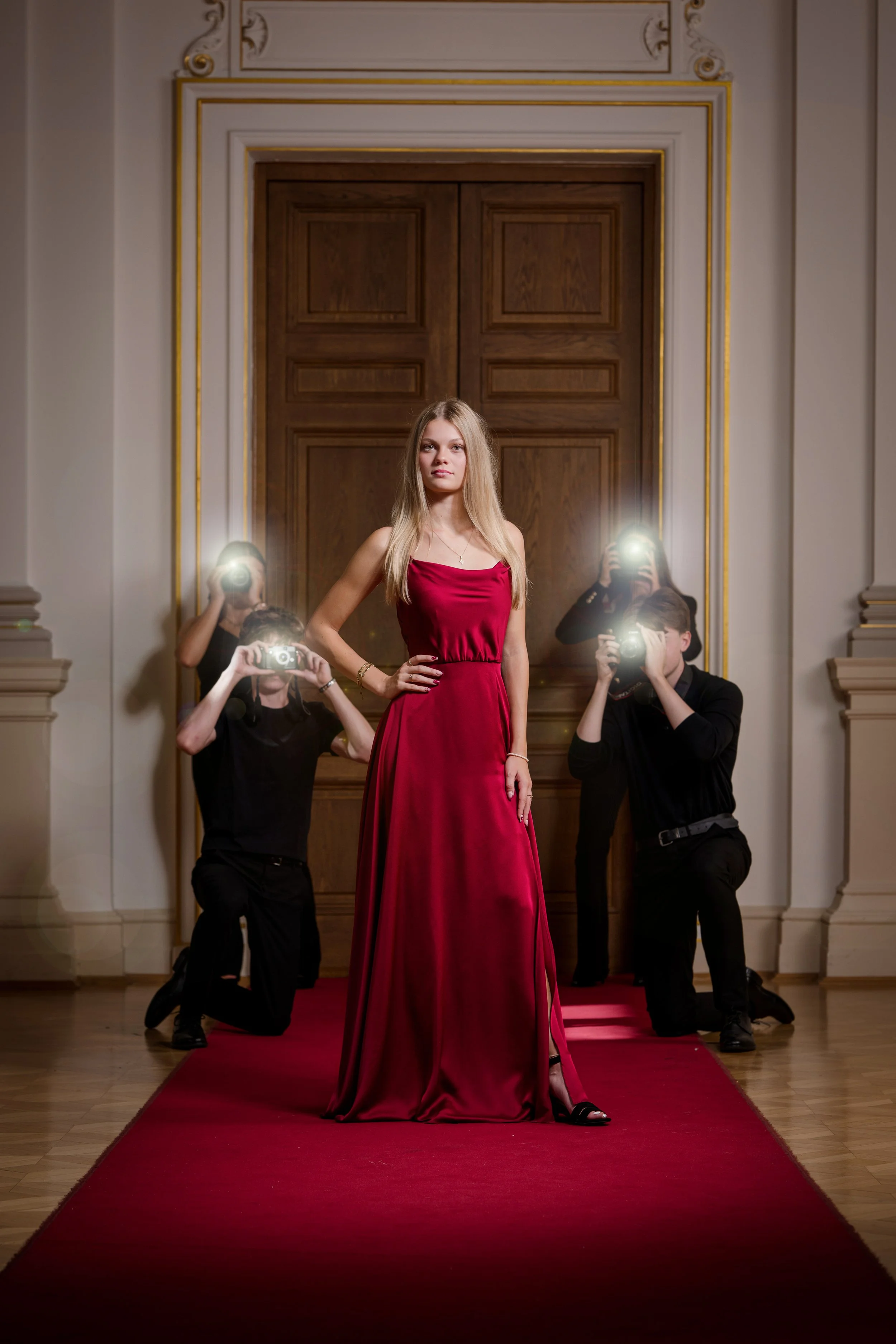Eine junge Frau in einem roten Abendkleid posiert auf einem roten Teppich, umgeben von Fotografen, die Fotos machen, in einem eleganten, dekorierten Raum.