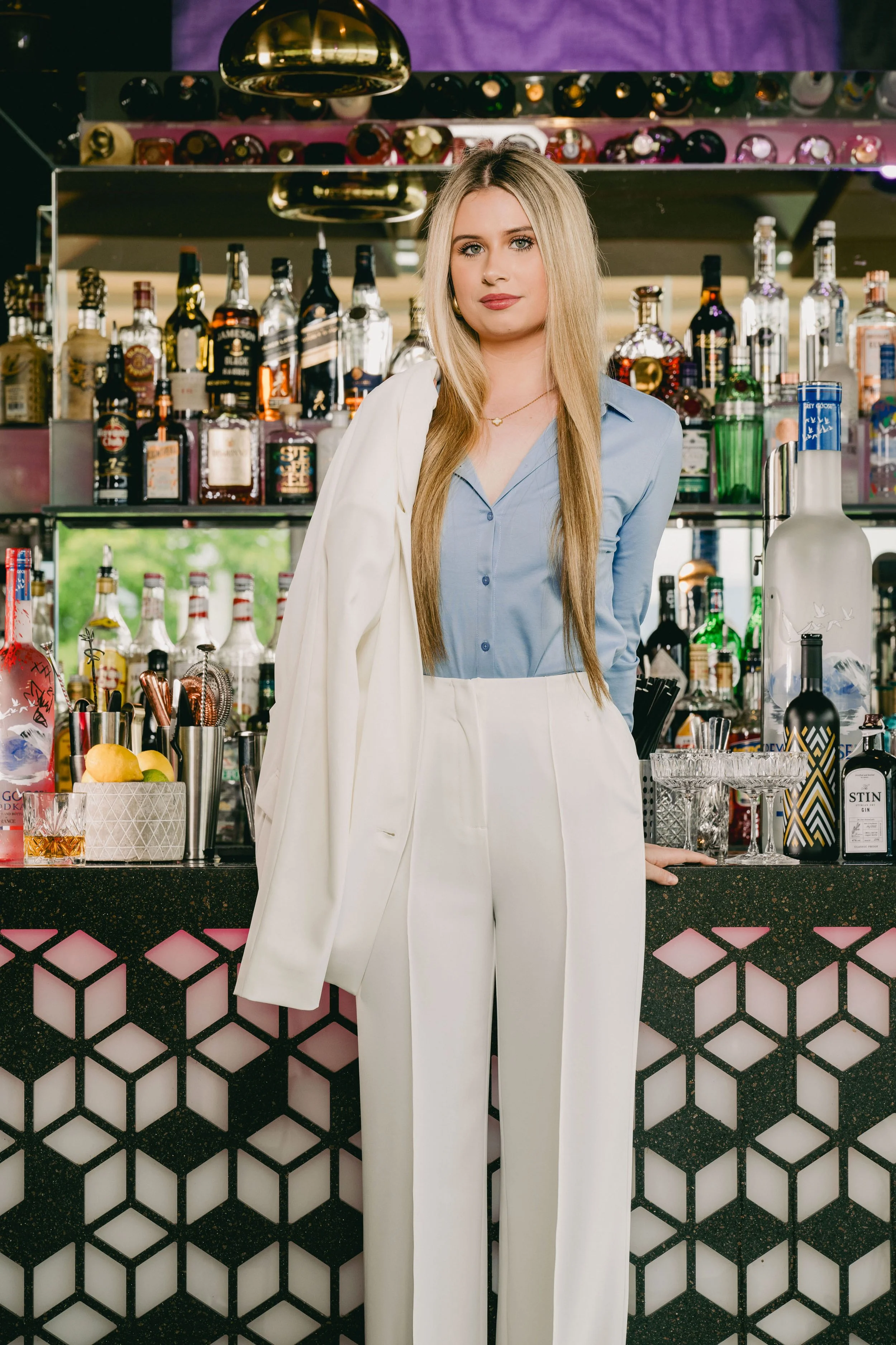 Eine junge Frau steht hinter einer Bar, umgeben von verschiedenen Alkoholflaschen, mit einem weißen Blazer über der Schulter, in einem modernen, gut beleuchteten Bar-Umfeld.