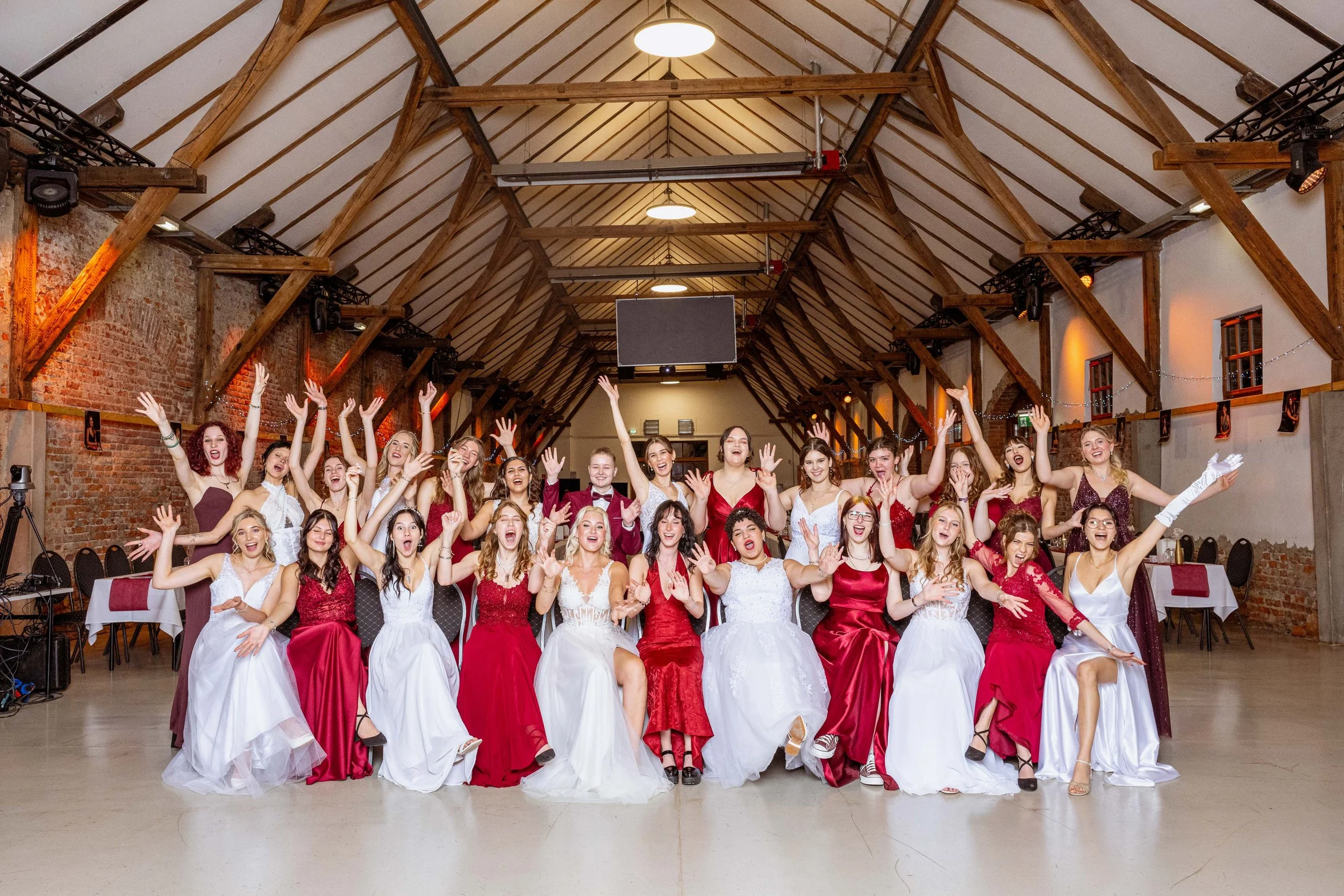 Eine Gruppe von jungen Frauen in eleganten Kleidern und einigen jungen Männern in Anzügen posieren in einem großen Veranstaltungsraum mit Holzdecke und roten Ziegelwänden, alle lachen und feiern.