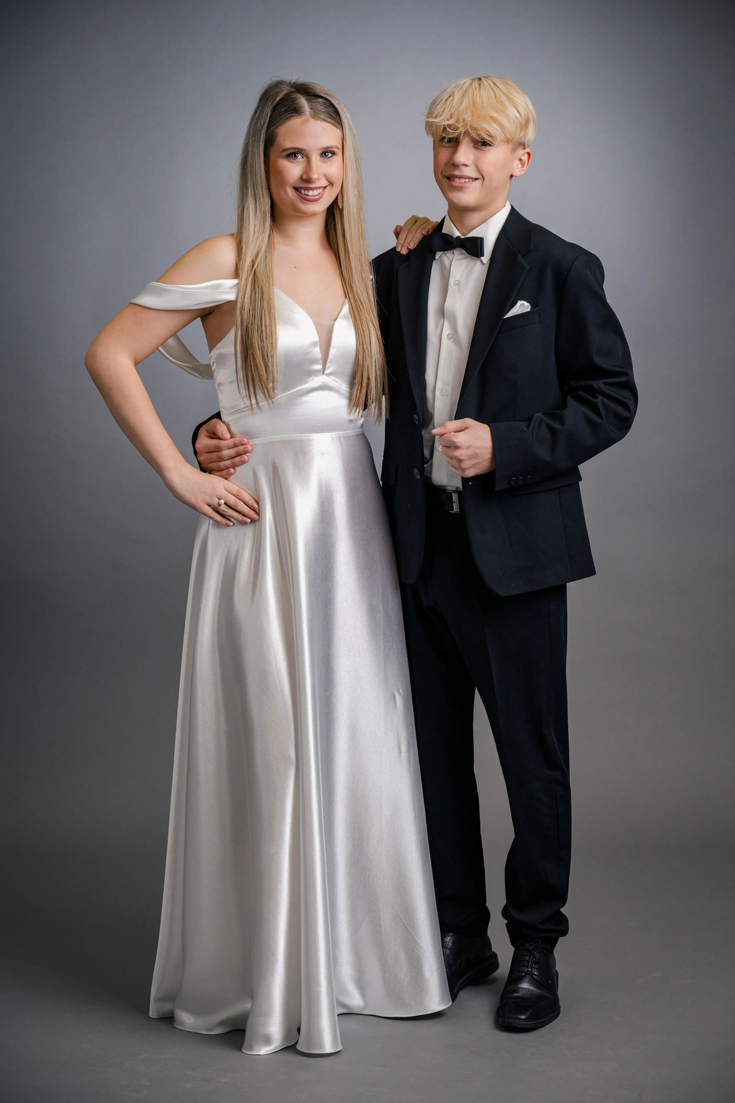 Ein junges Paar in eleganter Abendkleidung posiert vor grauem Hintergrund. Das Mädchen trägt ein weißes, großzügiges Abendkleid, der junge Mann einen schwarzen Anzug mit Fliege.