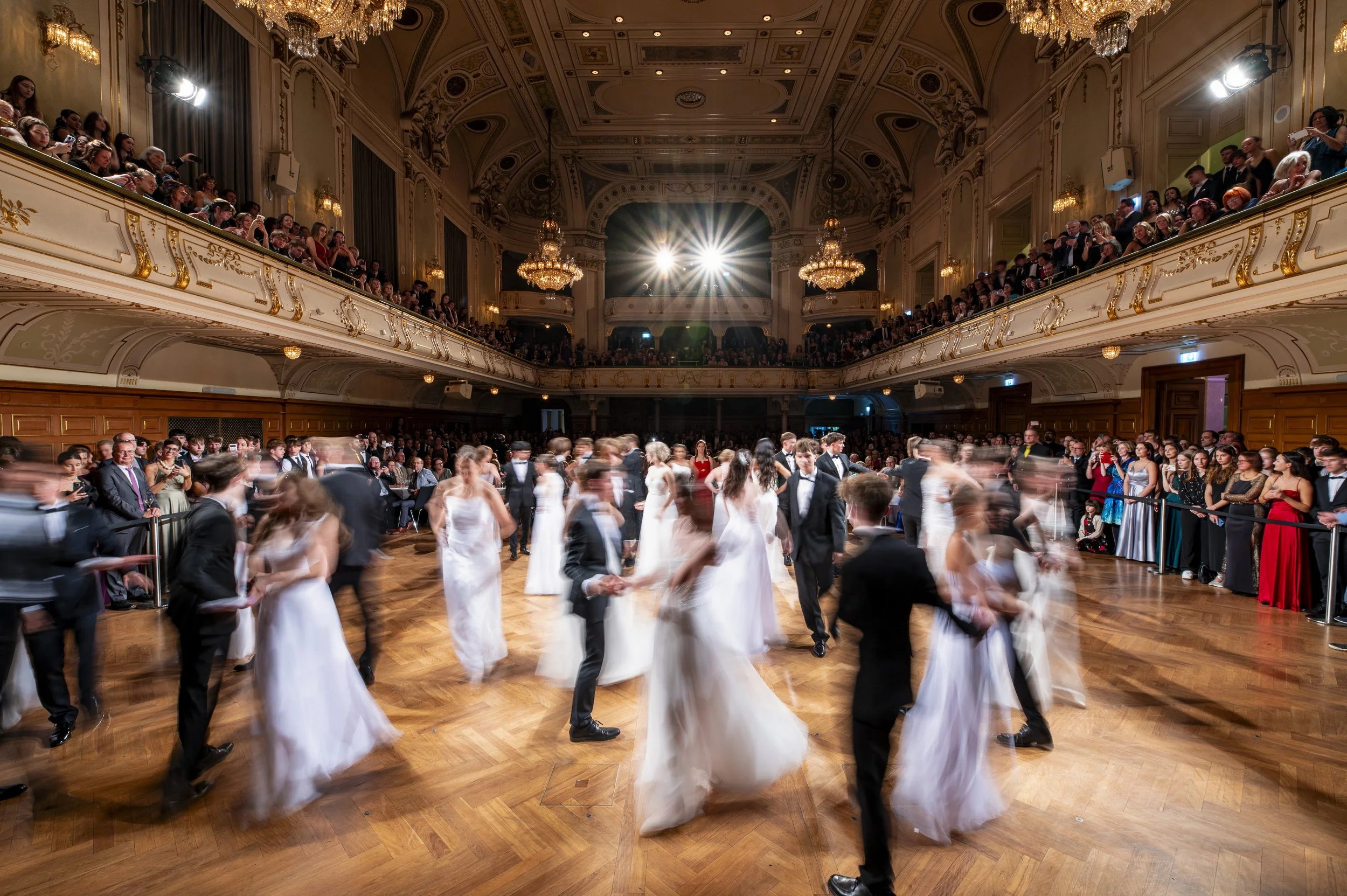 Tanzpaar mit Hochzeitskleidern und Smokings tanzt auf einer eleganten Tanzfläche in einem prunkvollen Saal mit Kronleuchtern, während Zuschauer zuschauen.