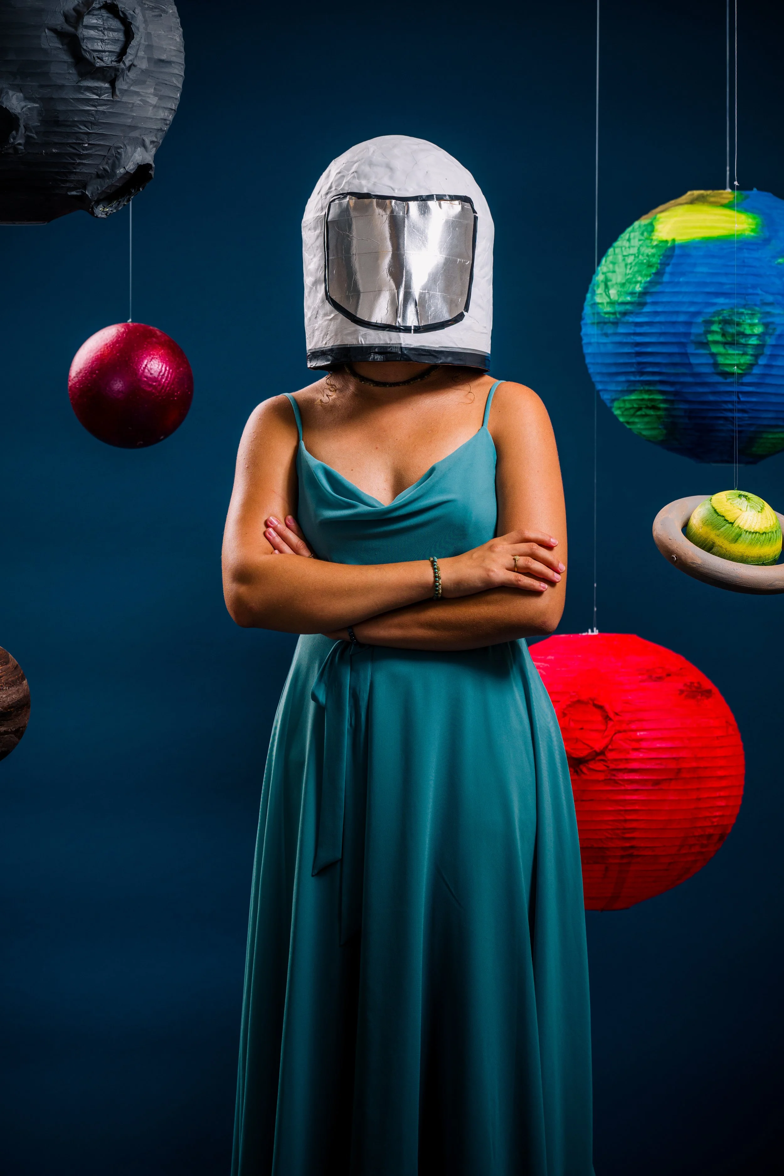 Eine Frau in einem blauen Kleid trägt eine Astronautenhelm-Kopie und steht vor dekorativen, schwebenden Lampions, die Planeten und die Erde darstellen.