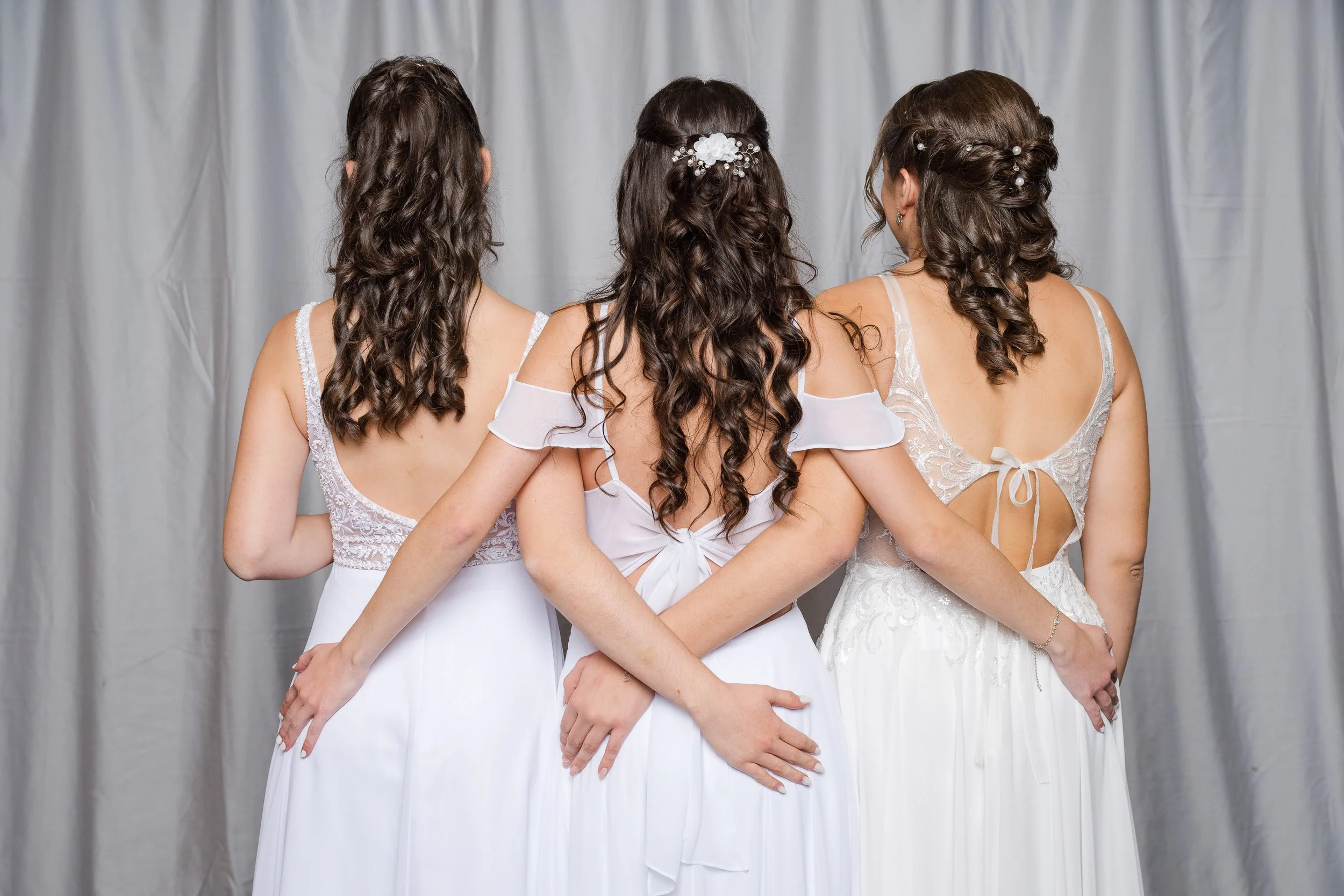 Drei Frauen in Brautkleidern stehen Rücken an Rücken vor einem weißen Vorhang, die Haare geflochten und mit Perlen und Blumen geschmückt.