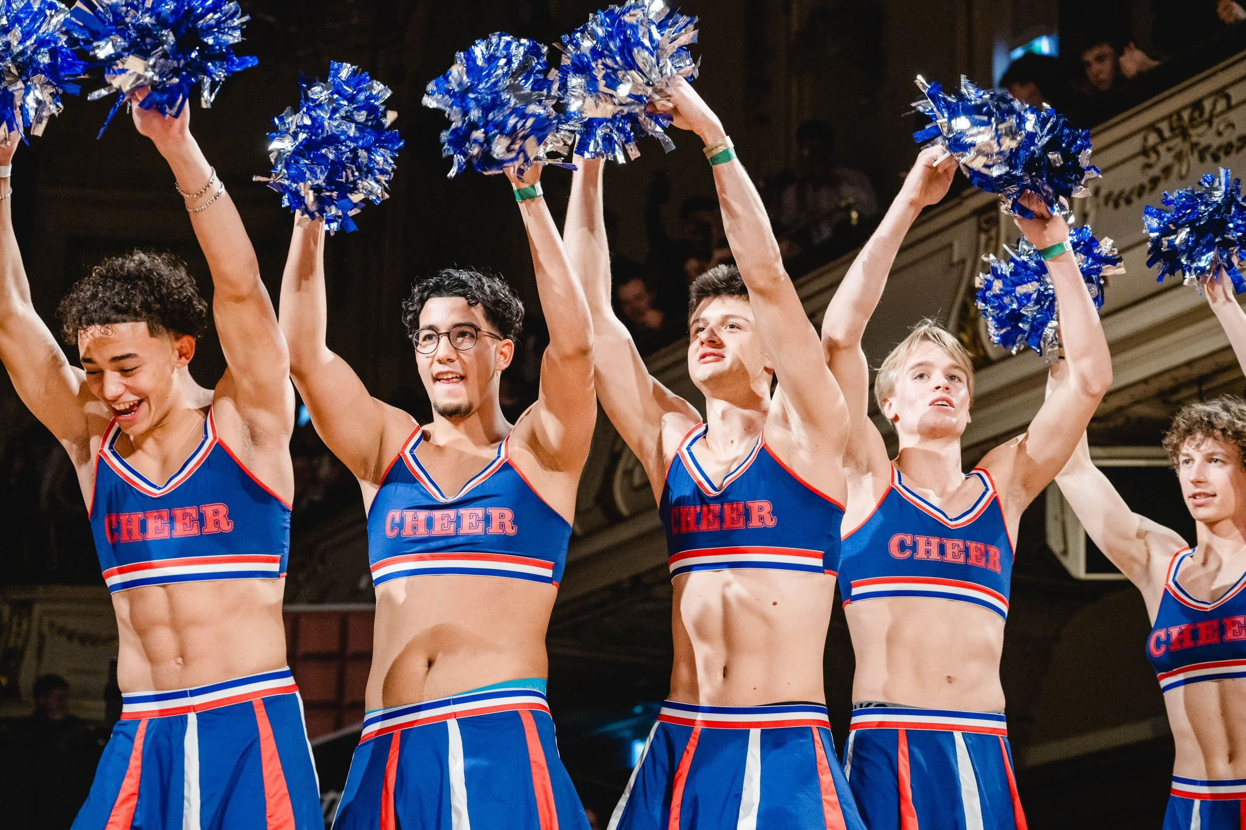 Gruppe von Cheerleadern in blauen, roten und weißen Uniformen mit der Aufschrift 'CHEER' auf ihrem Oberteil, die ihre Arme heben und mit blauen Pompons fröhlich feiern.