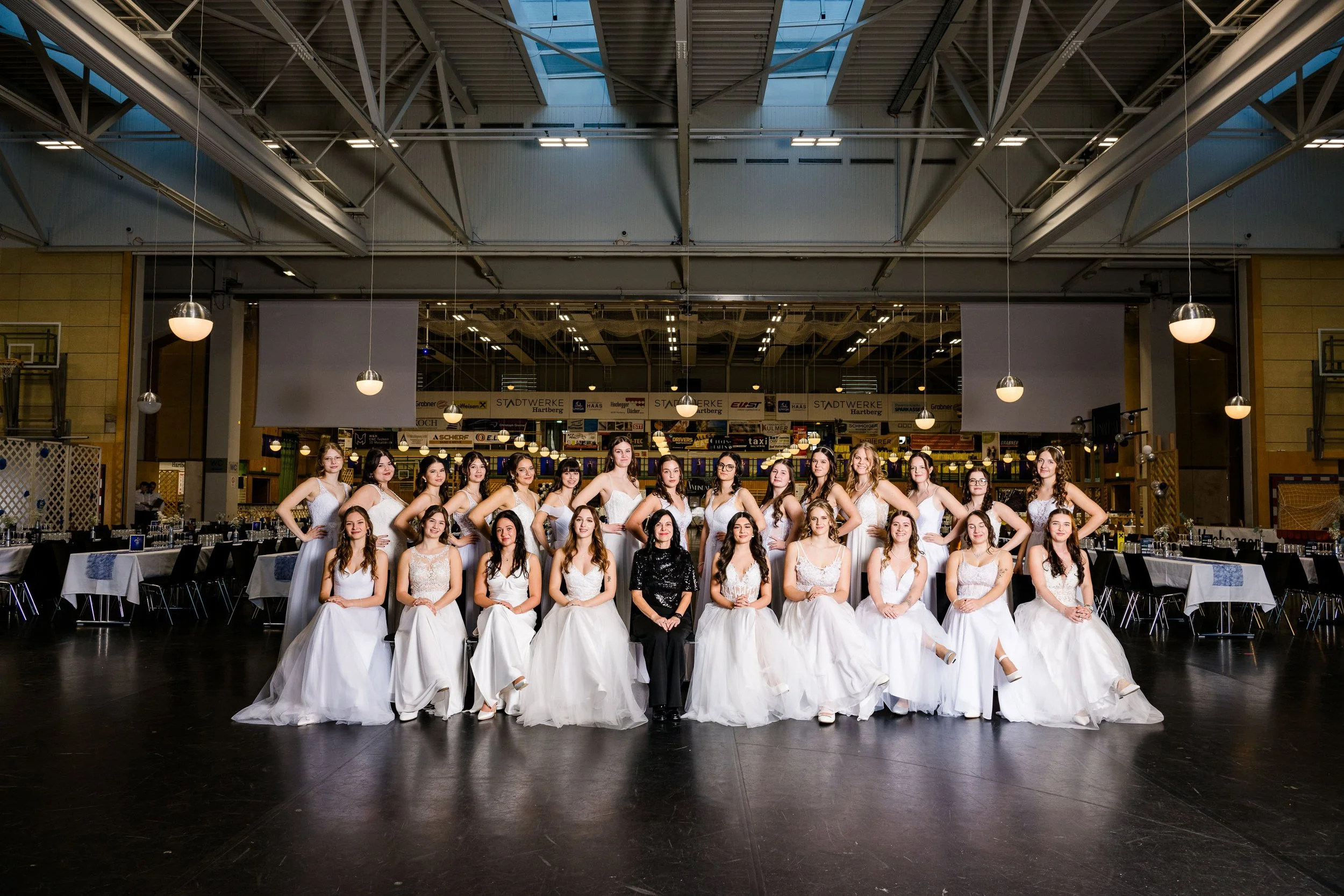 Gruppe junger Frauen in weißen Kleidern, eine Frau in schwarzem Kleid, in einem Saal für eine Gala oder Hochzeit.
