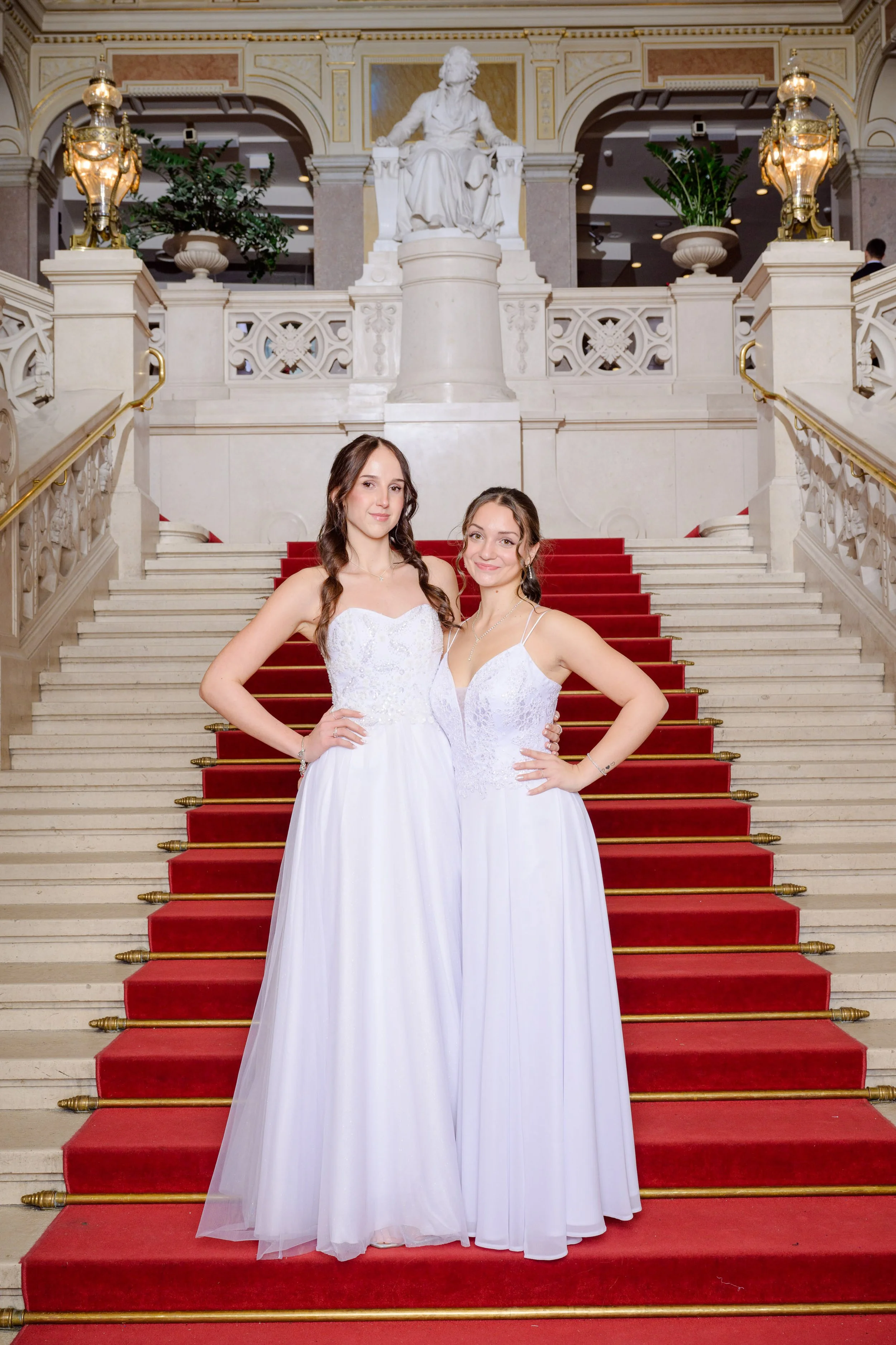 Zwei junge Frauen in weißen Brautkleidern posieren auf einer prächtigen Treppe mit rotem Teppich und goldenen Handläufen, im Hintergrund eine weiße Statue und elegante Verzierungen im eleganten Innenraum.