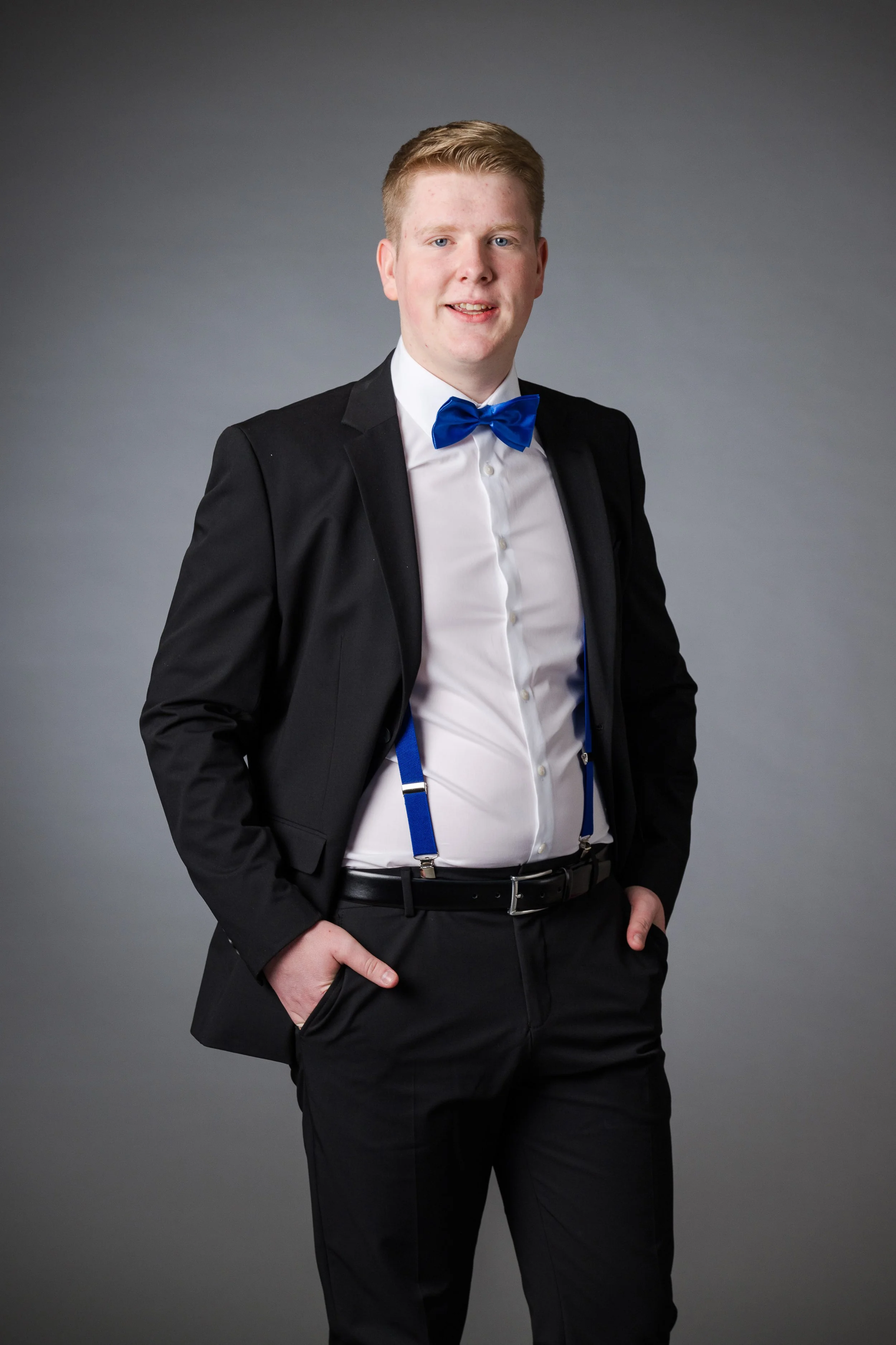 Ein junger Mann in schwarzer Anzugjacke mit weißem Hemd, blauer Fliege und Hosenträgern, pose with hands in den Taschen vor grauem Hintergrund.