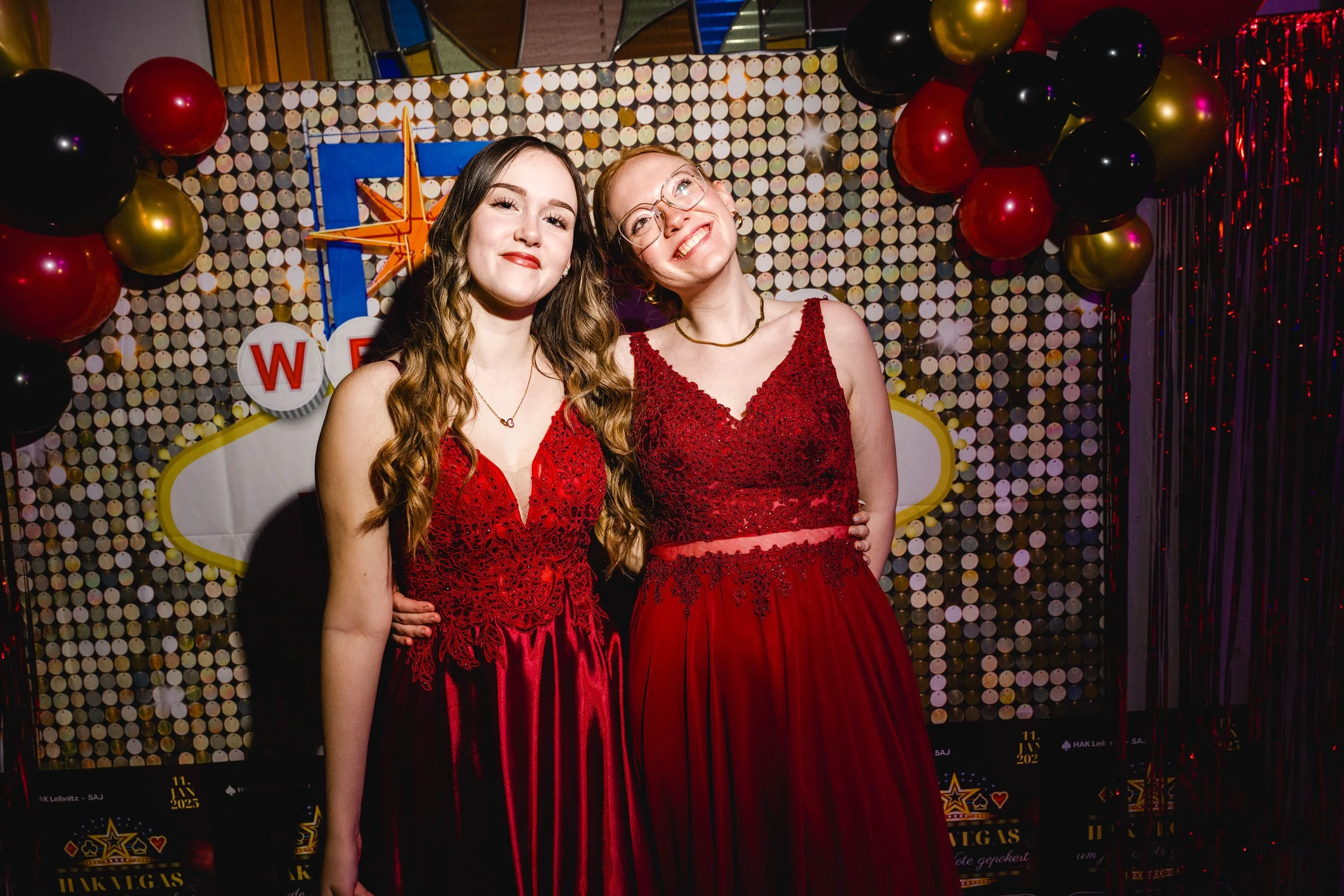 Zwei lachende junge Frauen in roten, eleganten Kleidern auf einer Party, im Hintergrund Luftballons und eine festliche Dekoration.
