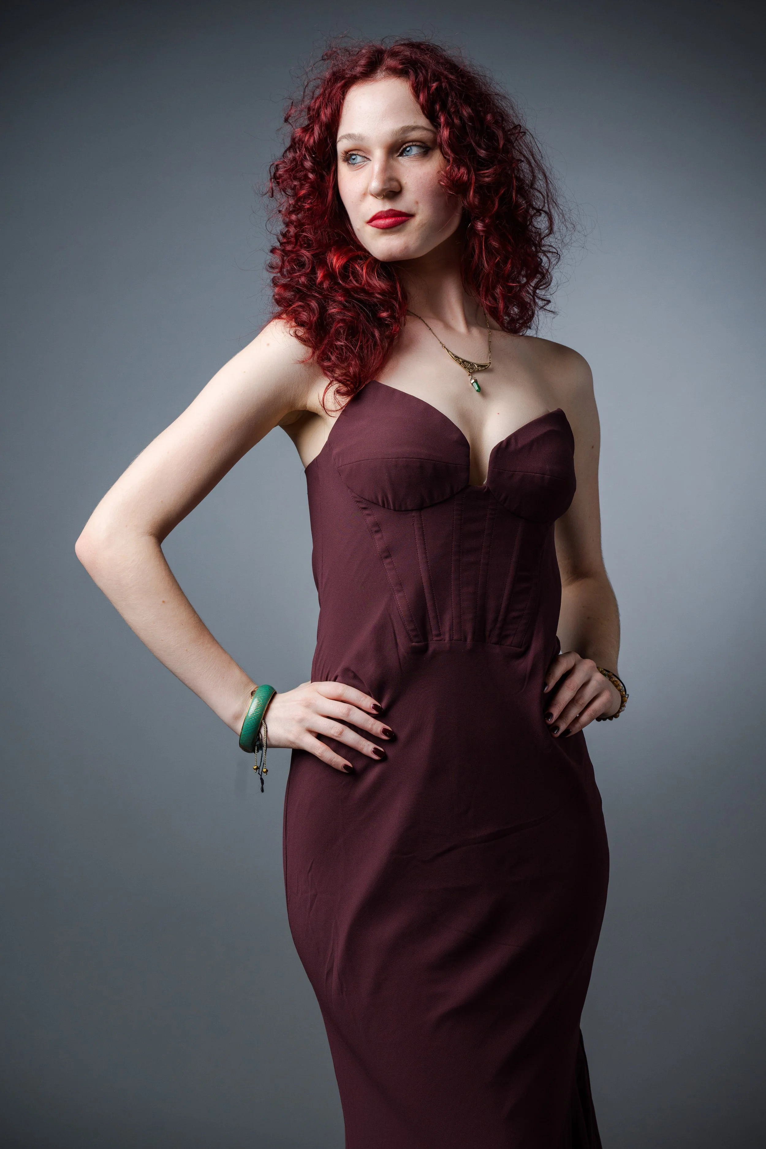 Eine Frau mit roten, lockigen Haaren trägt ein dunkles rotes, körperbetontes Kleid. Sie steht vor einem grauen Hintergrund und schaut seitlich, mit Schmuck und ihrer Hand auf der Hüfte.