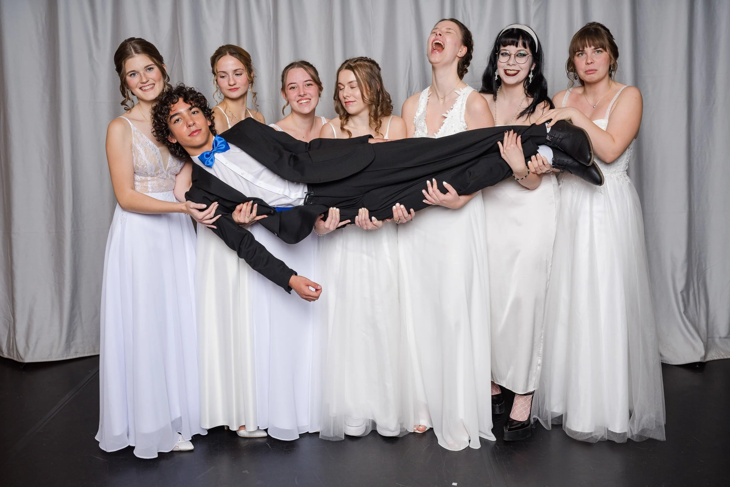 Eine Gruppe von acht Frauen in weißen Kleidern halten einen männlichen Jungen in einem schwarzen Anzug mit blauer Fliege hoch. Alle lachen und posieren vor einem grauen Hintergrund.
