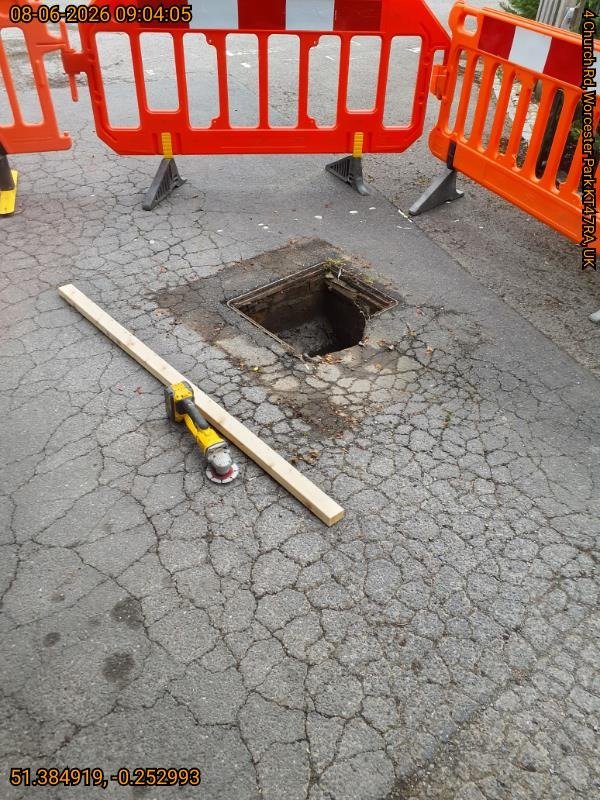 Open manhole with its broken cover removed in a Worcester Park restaurant car park, secured with safety barriers while awaiting a replacement
