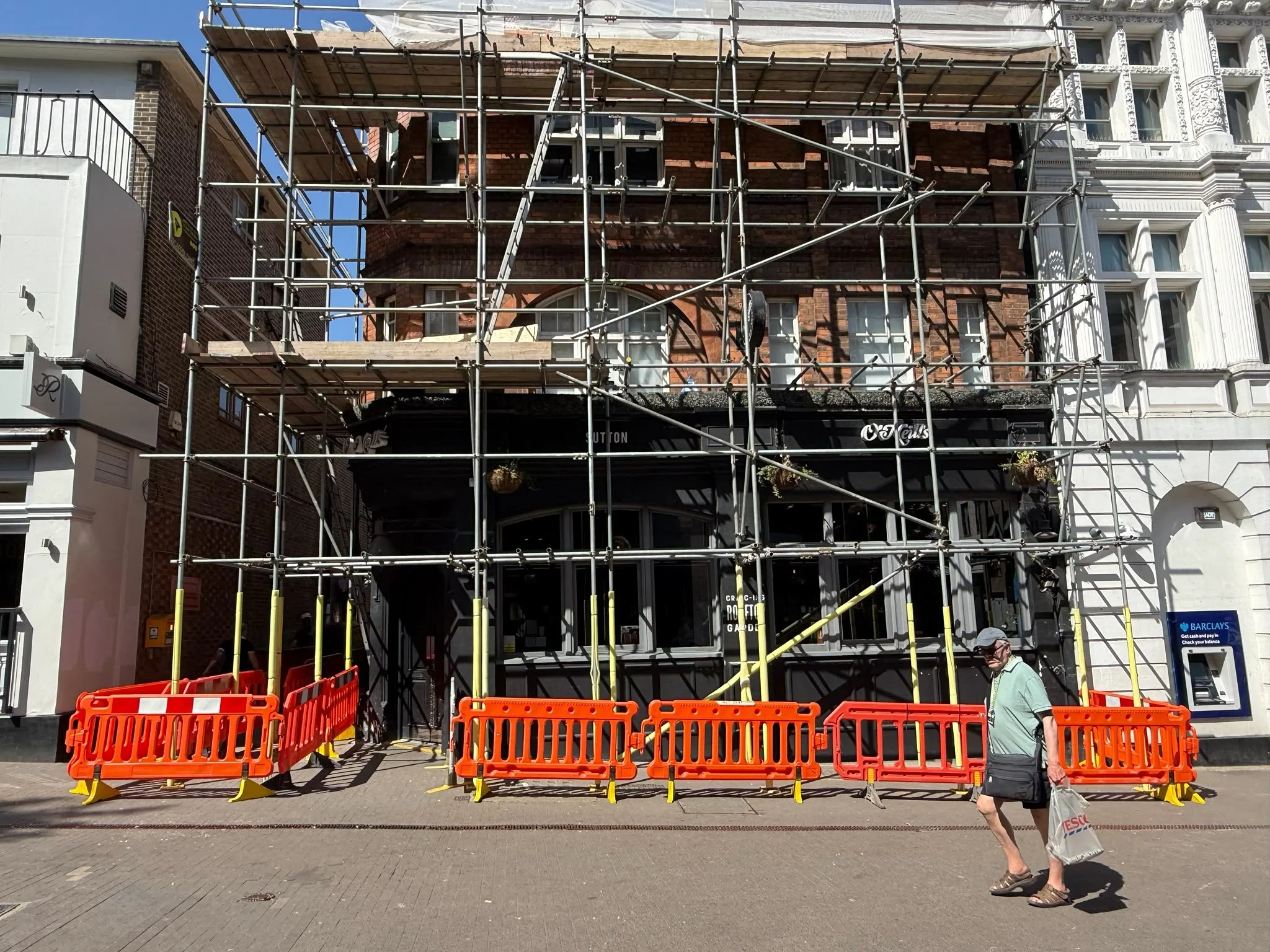 Scaffolding and safety barriers at street level during the parapet wall repair on a Sutton high street, with the pavement kept open for pedestrians