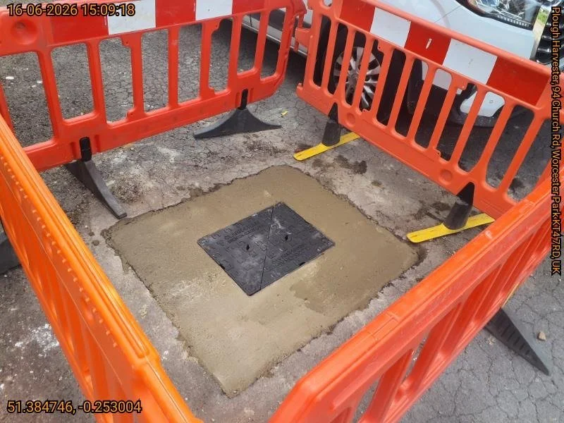New heavy-duty manhole cover bedded in fresh concrete in the Worcester Park car park, surrounded by safety barriers while it cures