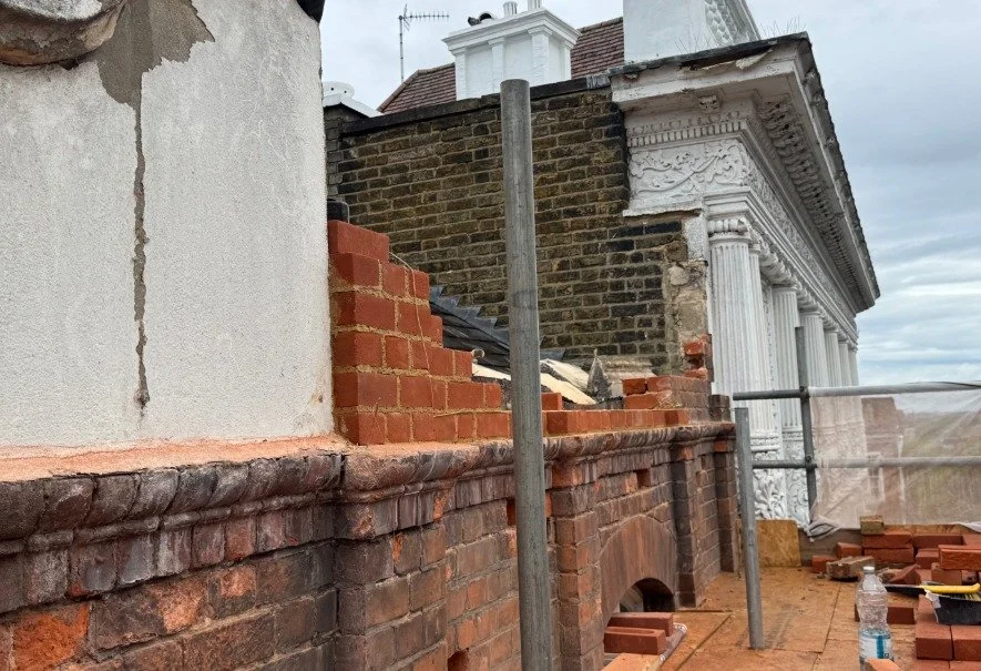 Partially rebuilt parapet wall in Sutton showing new red brickwork against the original wall, with scaffolding poles and a period building behind
