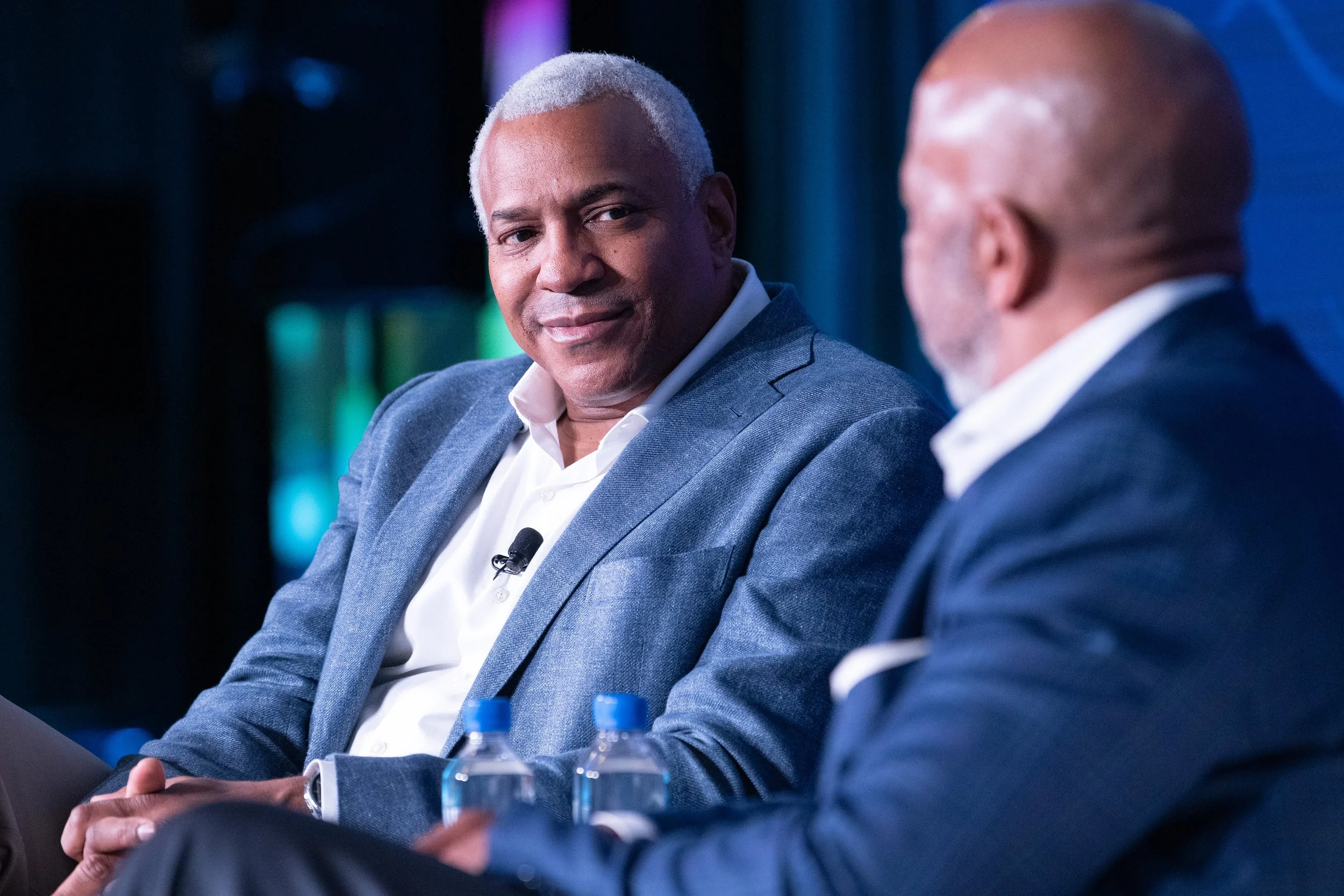 Two men in business suits, sitting and facing each other, engaged in conversation. The man on the left has gray hair, is smiling slightly, and looking at the man on the right, who is balding with white hair and turned to his left.