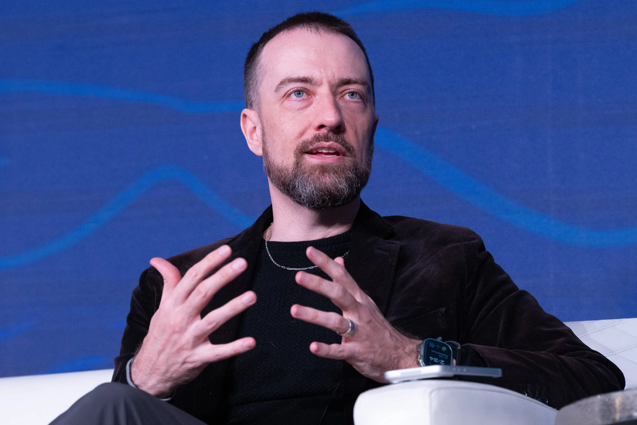 A man with a beard and short hair speaking during a panel or discussion, gesturing with his hands. He is wearing a black blazer, a black shirt, a silver necklace, and a smartwatch. The background is blue with abstract lines.