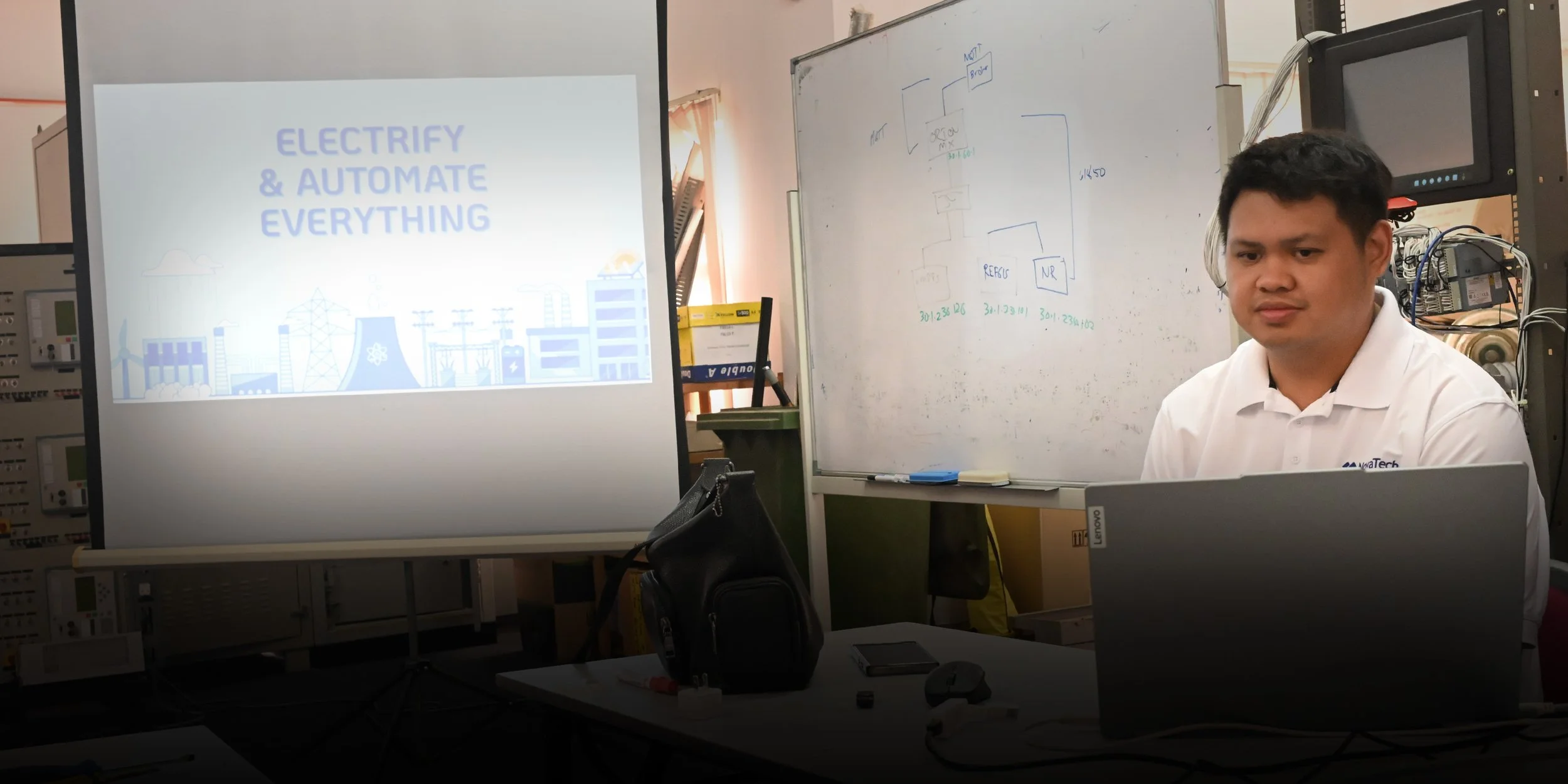 Young man giving a presentation with a microphone, standing next to a laptop in a conference room with a screen displaying logos.