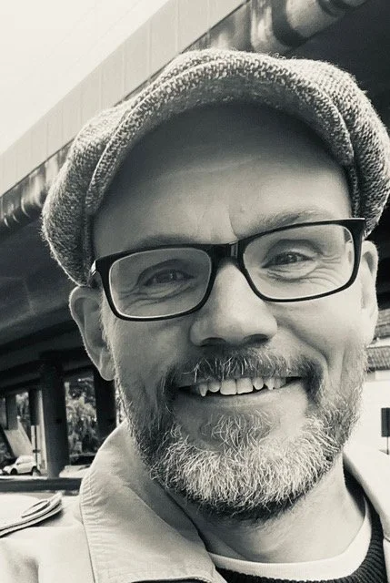 Mann med skjegg iført rutete jakke og caps, poserer ved et vindu.