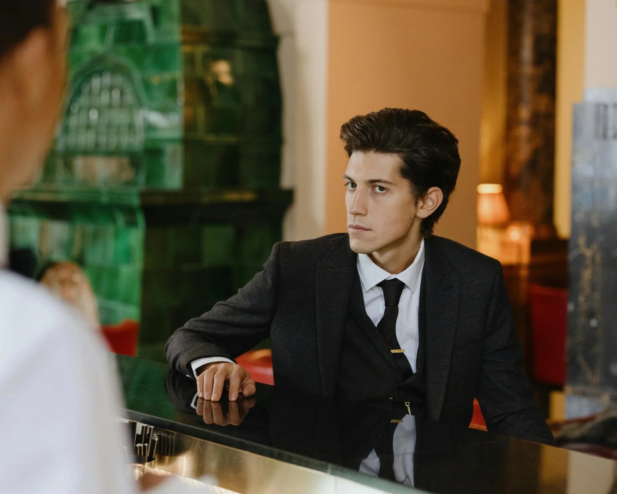 Un homme jeune en costume noir, assis à un piano dans un lieu décoré avec des lampes et des meubles chaleureux, regarde fixement une personne hors champ.