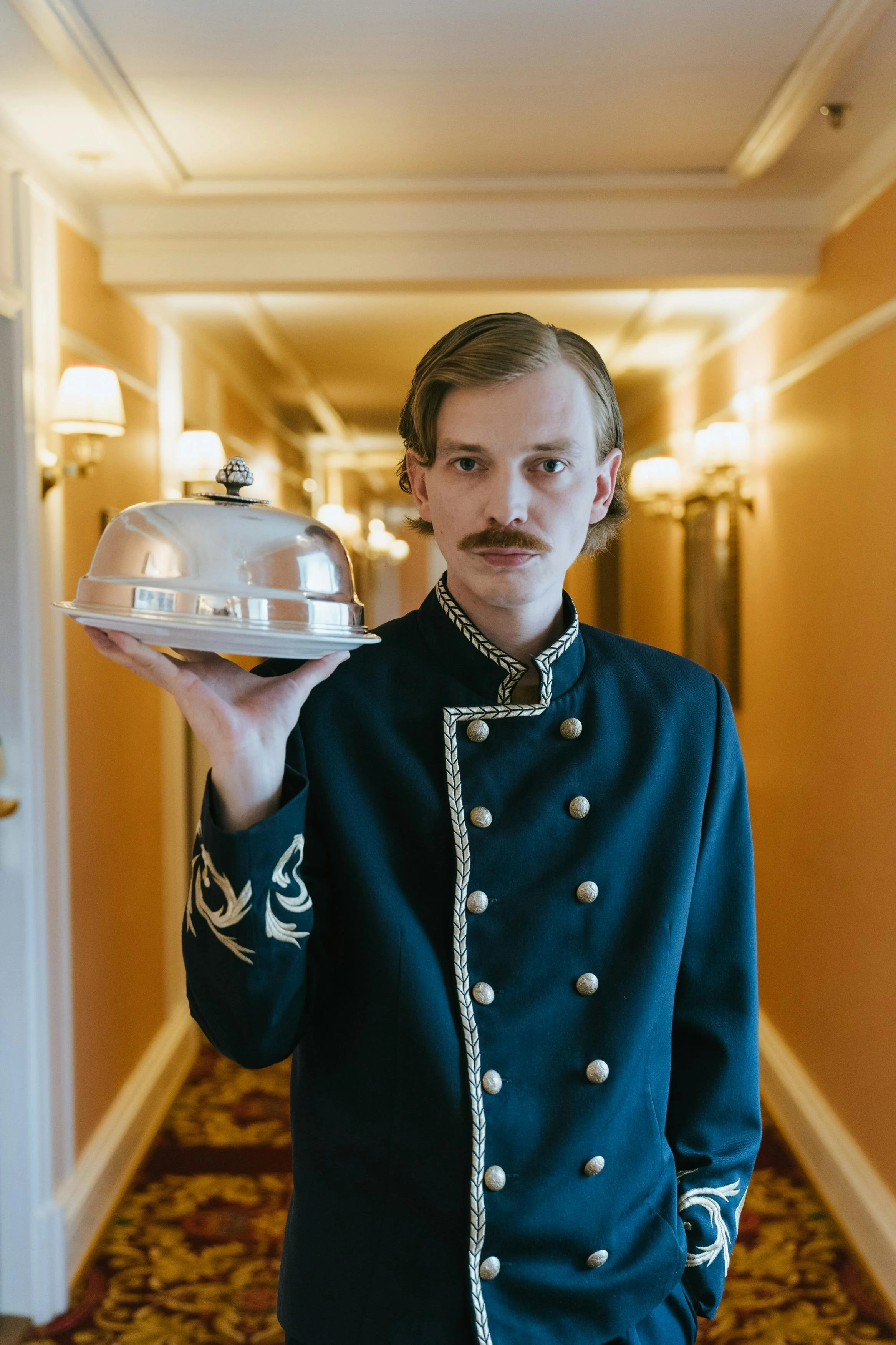 Un serveur en uniforme noir avec des boutons argentés et des détails ornementaux blancs dans un couloir lumineux d'hôtel, tenant un plateau avec une cloche en aluminium