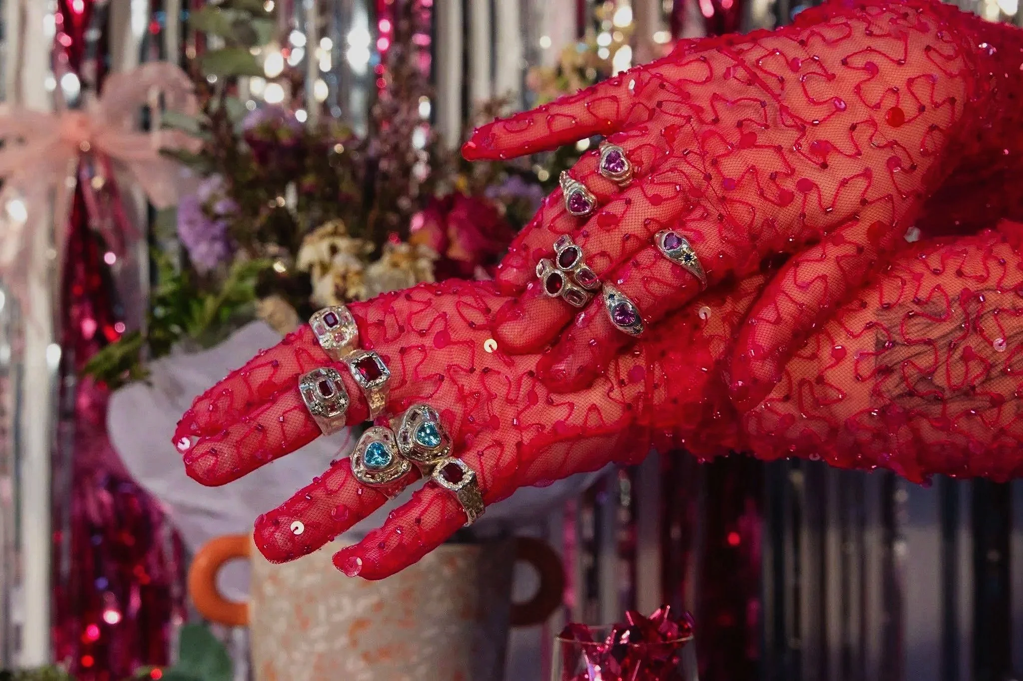 A hand wearing red, lace gloves adorned with sequins, with multiple silver rings featuring colorful gemstones and heart-shaped designs, situated in front of a backdrop with flowers and shiny decorations.