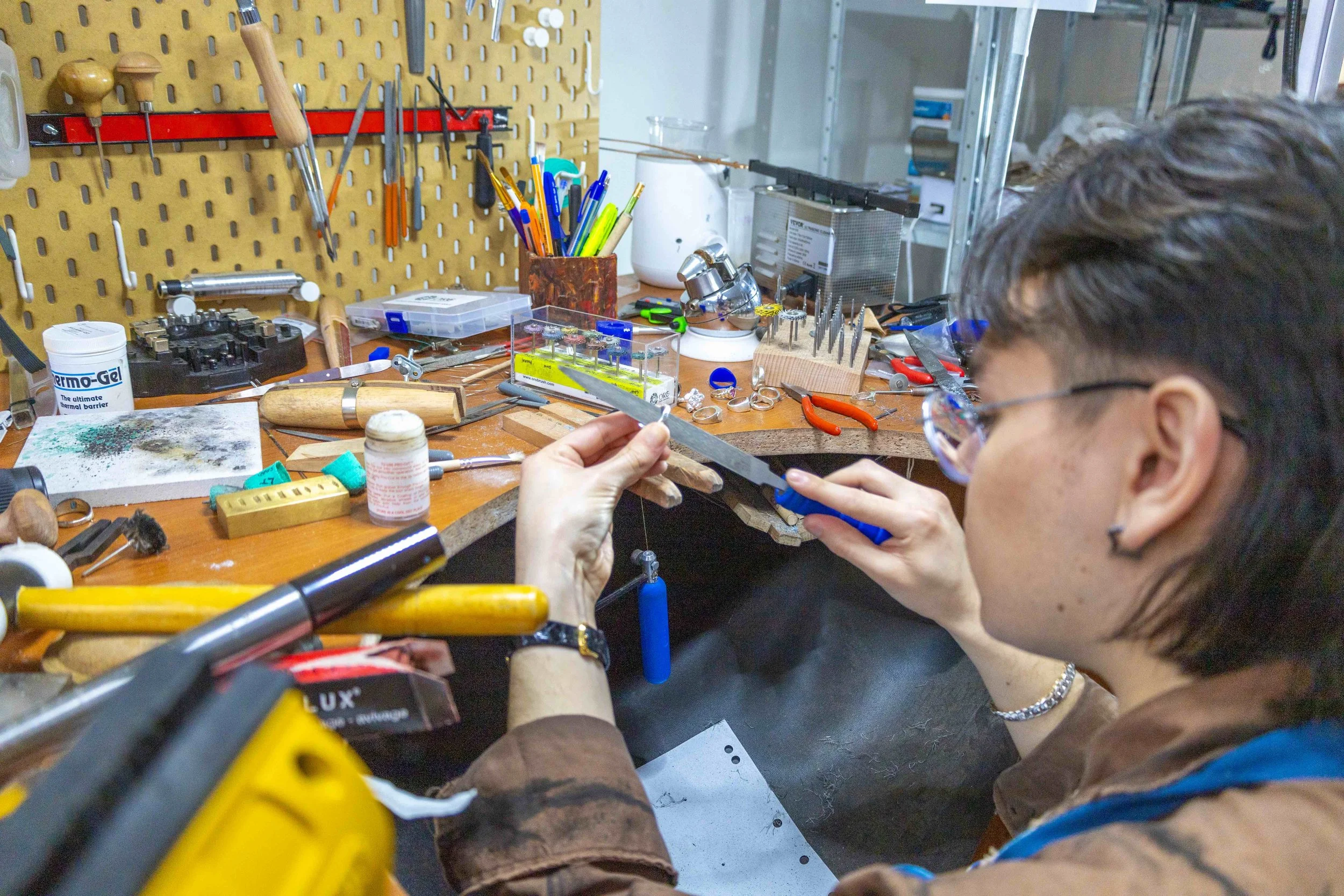 A person working on a jewelry piece in a cluttered jewelry workshop, surrounded by tools, pliers, blades, and supplies on a workbench.