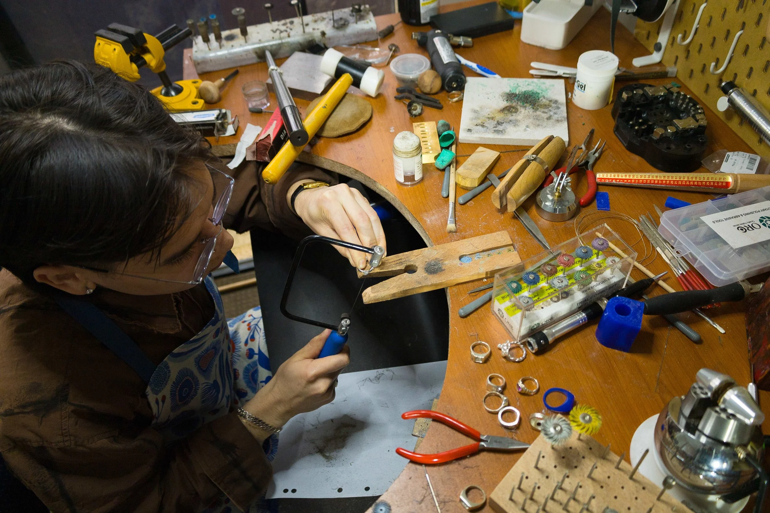 A woman working at a cluttered jewelry repair or creation station with various tools, materials, and jewelry pieces on the workbench.