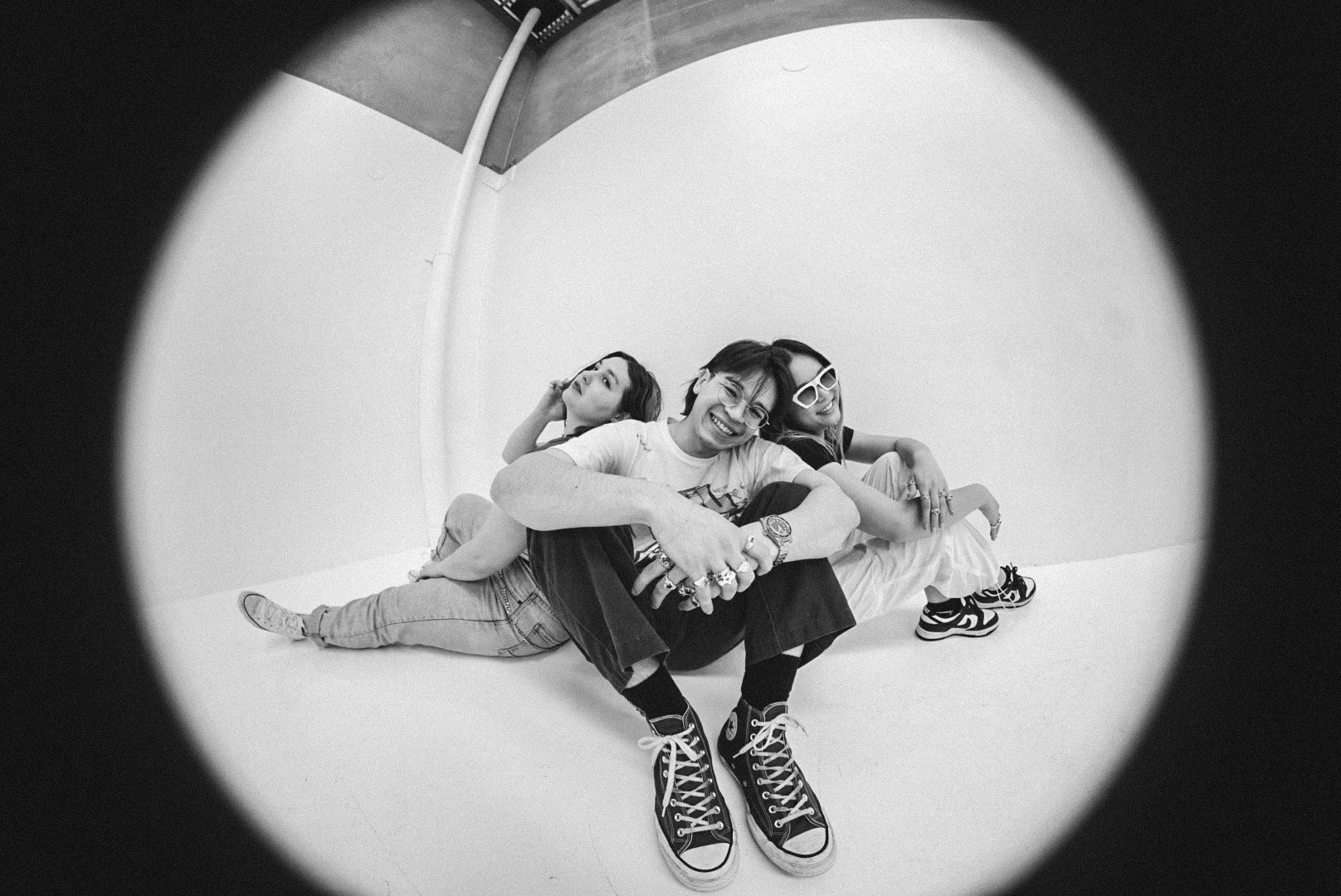 Three women sitting on the floor, smiling and posing for a photo, with a camera fisheye lens effect. One woman in the front with glasses and sneakers, smiling widely. Two women behind her, one with sunglasses and the other in a casual pose with their head resting on their hand.