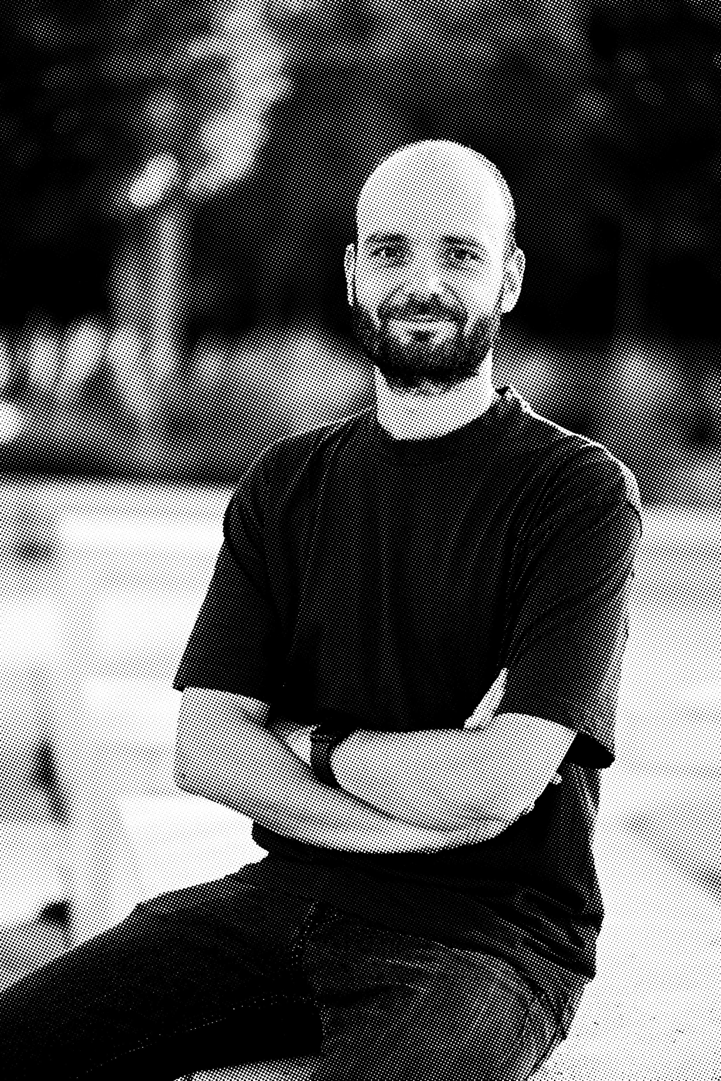 A man with a bald head, beard, and mustache sitting with his arms crossed and smiling. He is wearing a short-sleeved, dark shirt and jeans, in a studio setting with a dark background.