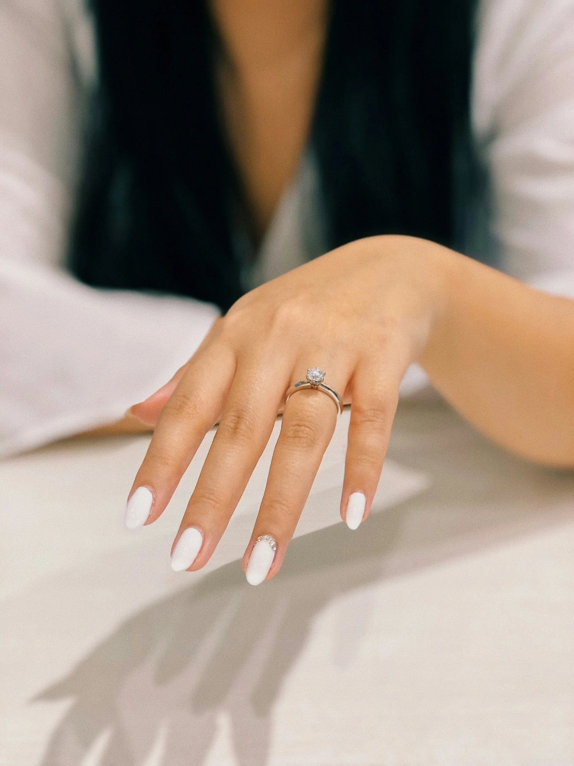 A woman's hand with white painted nails displaying a diamond engagement ring on her ring finger.