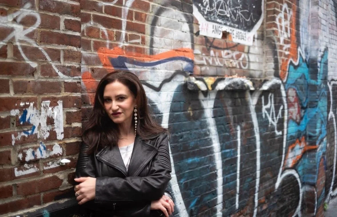 A woman with dark hair, wearing a black leather jacket and pearl earrings, standing in front of a brick wall covered in colorful graffiti art.