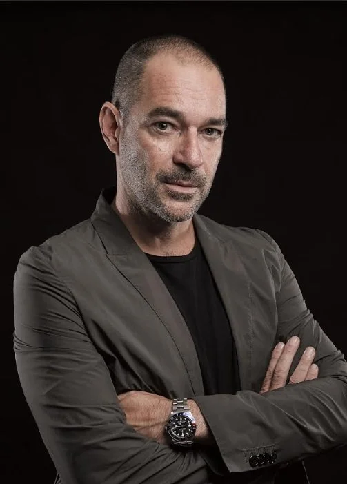 A man with a shaved head and beard, wearing a dark jacket over a black shirt, with arms crossed, against a black background.