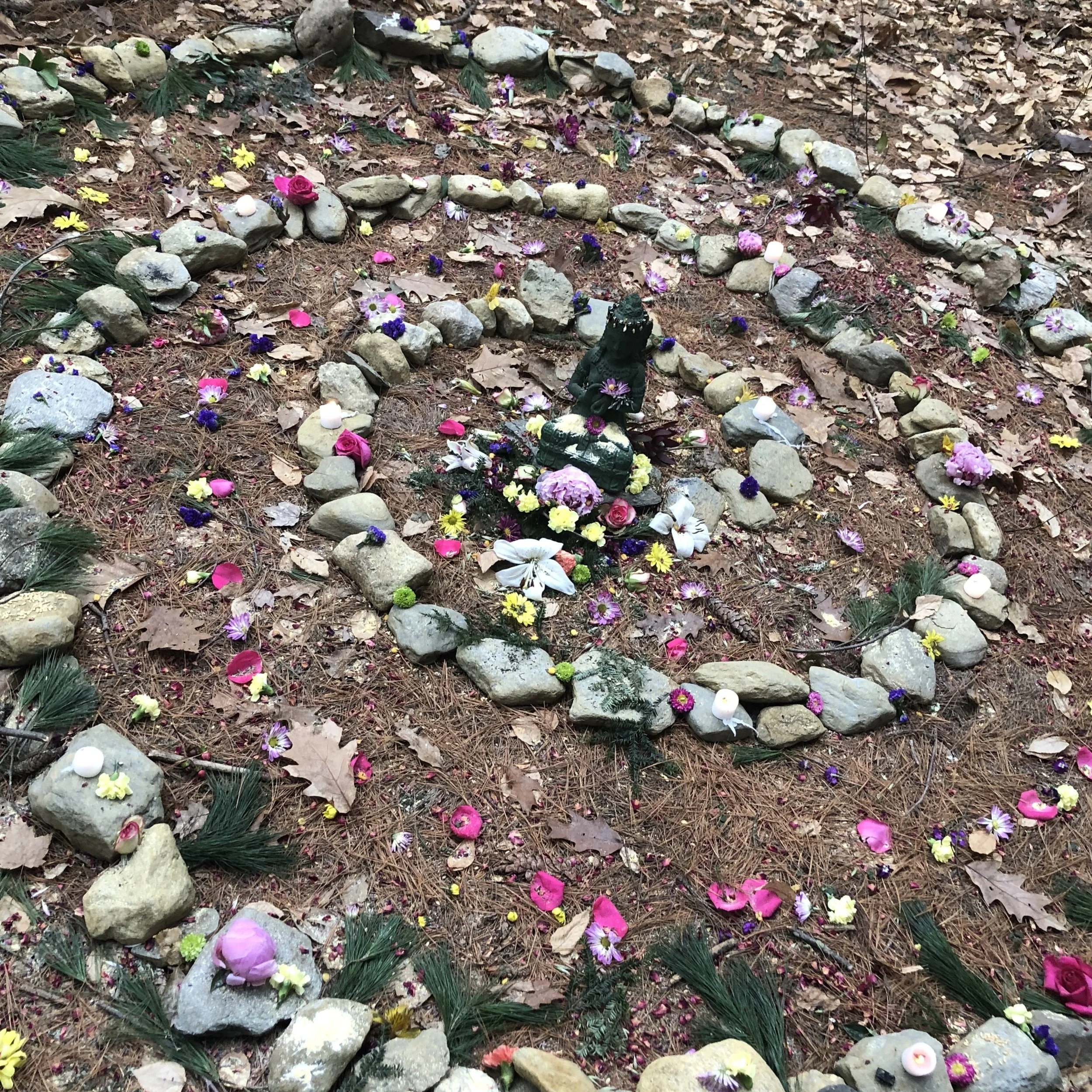 small stones lie in spiral pattern on bed of brown pine needles. Center of spiral sits a green statue sitting in lotus position. The spiral is decorated with various flower petal colors and freshly picked green pine boughs.