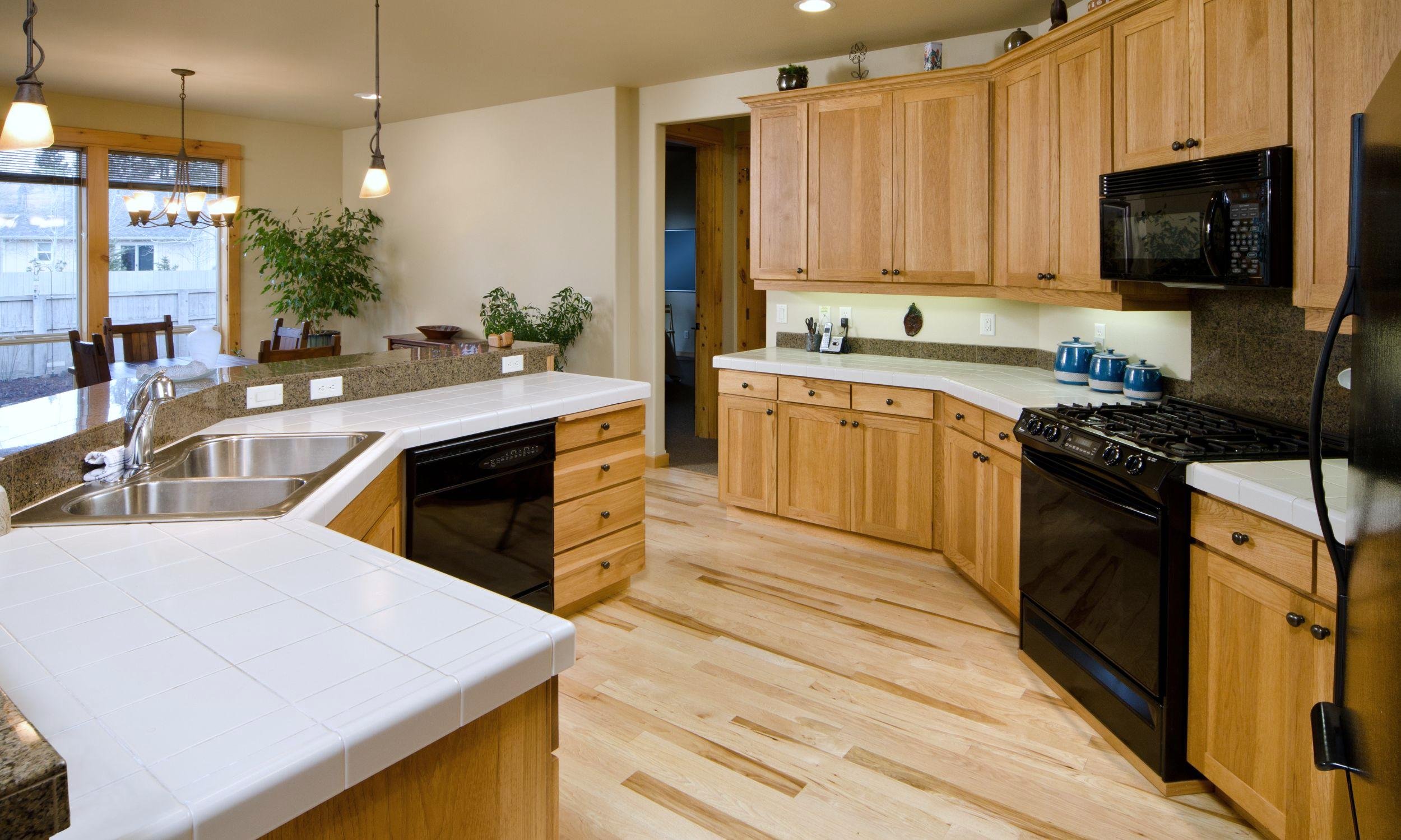 Kitchen with light wood floor and matching cabinets. White tile counter tops and black appliances. This is an example of house cleaning done by Scrubbles Cleaning Service.